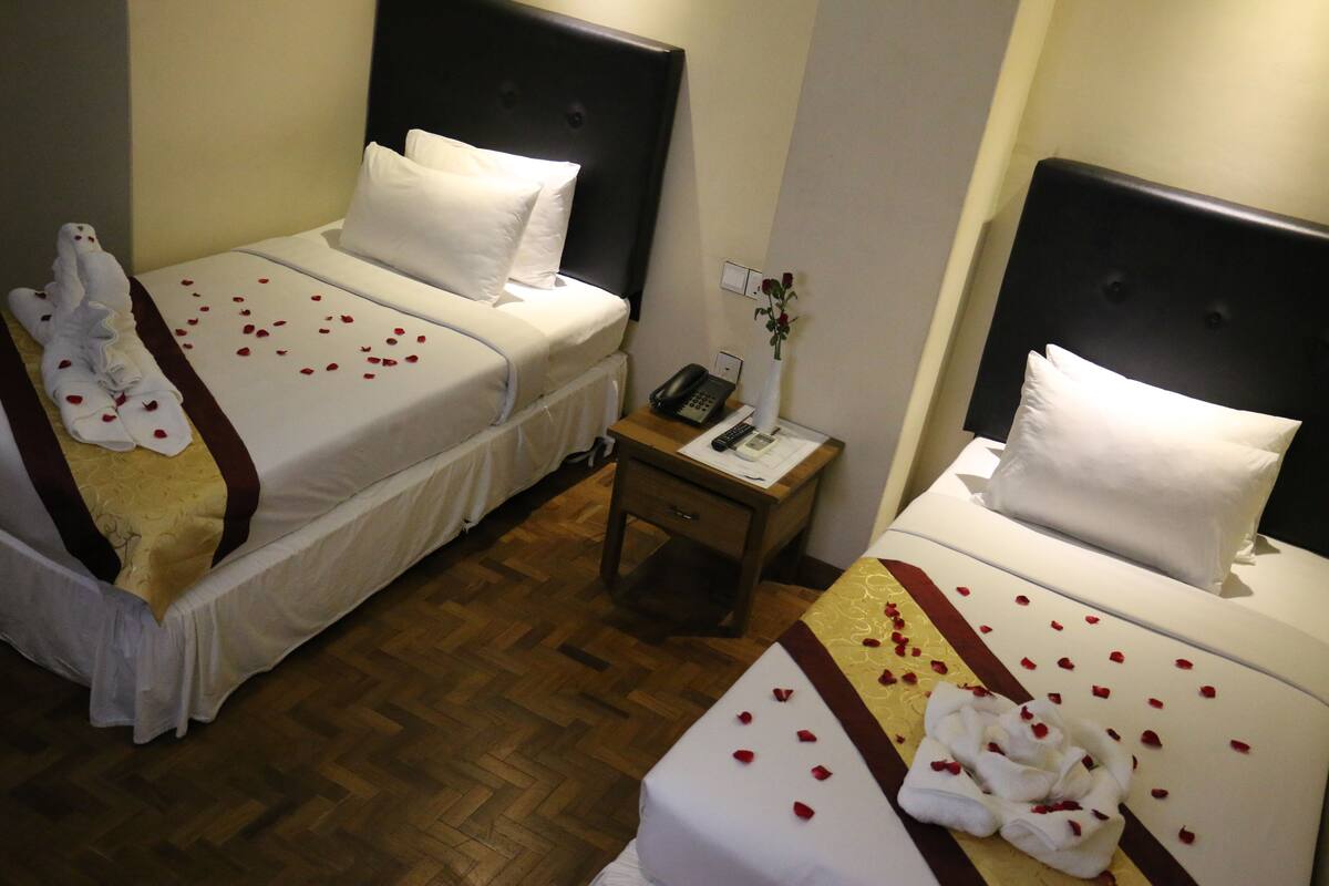 A hotel room with two beds, each with white linens and rose petals scattered on the beds. There is a nightstand with a phone and a vase with a single flower between the beds.