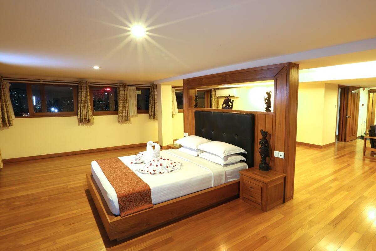 A spacious bedroom with a large bed, wooden floor, and a view of the city at night.