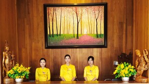 A reception desk with three staff members in yellow uniforms, wooden paneling, a framed painting of a forest, and decorative statues. - Queen's Park Hotel (Yangon)