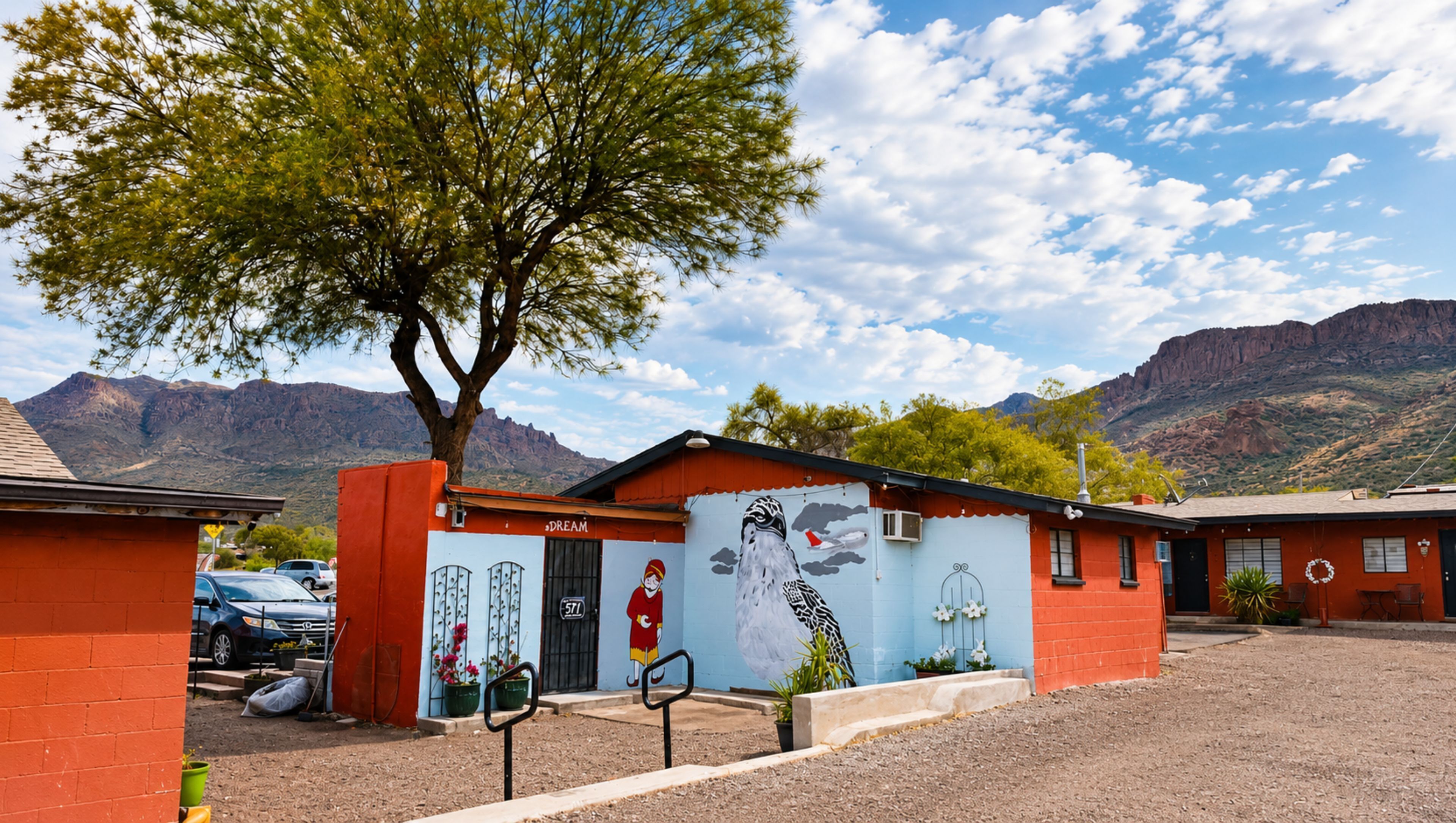 Un edificio con un mural, un árbol en primer plano y montañas al fondo.