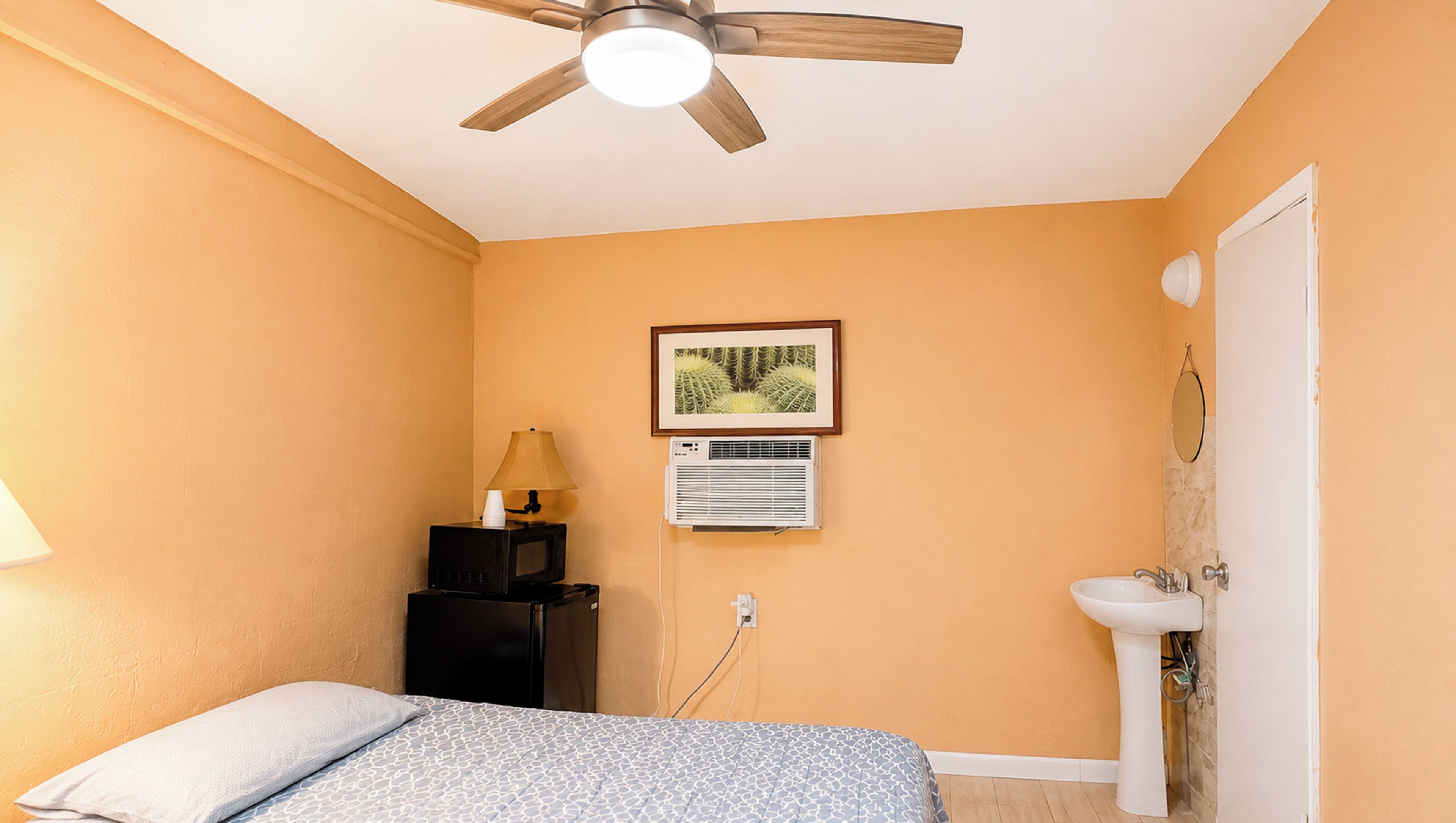 A bedroom with a bed, a ceiling fan, a framed picture, an air conditioning unit, and a bathroom with a sink.