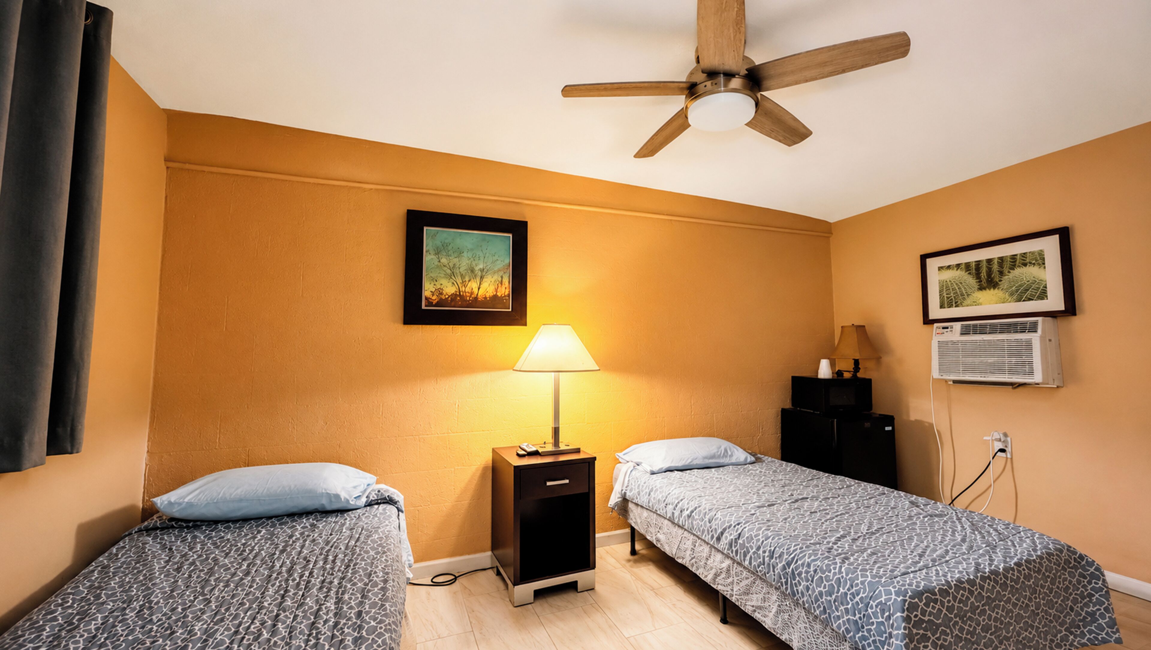 Una habitación con dos camas individuales, ventilador de techo, mesita de noche con lámpara, aire acondicionado y dos cuadros en la pared.