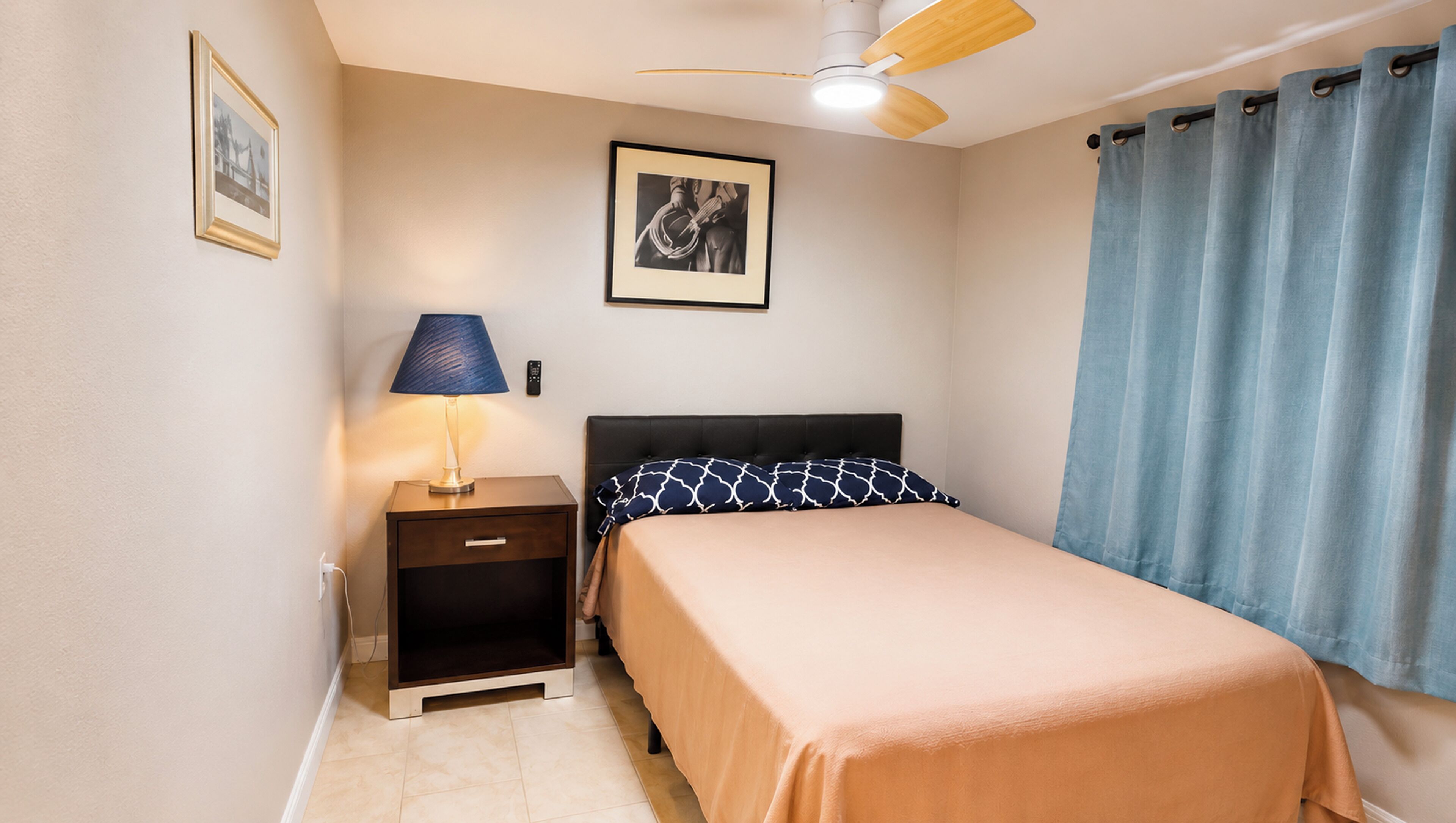 A bedroom with a bed, nightstand, lamp, ceiling fan, and a framed picture on the wall.