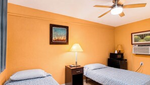 A hotel room with two beds, a ceiling fan, a nightstand with a lamp, a framed picture, and an air conditioning unit.