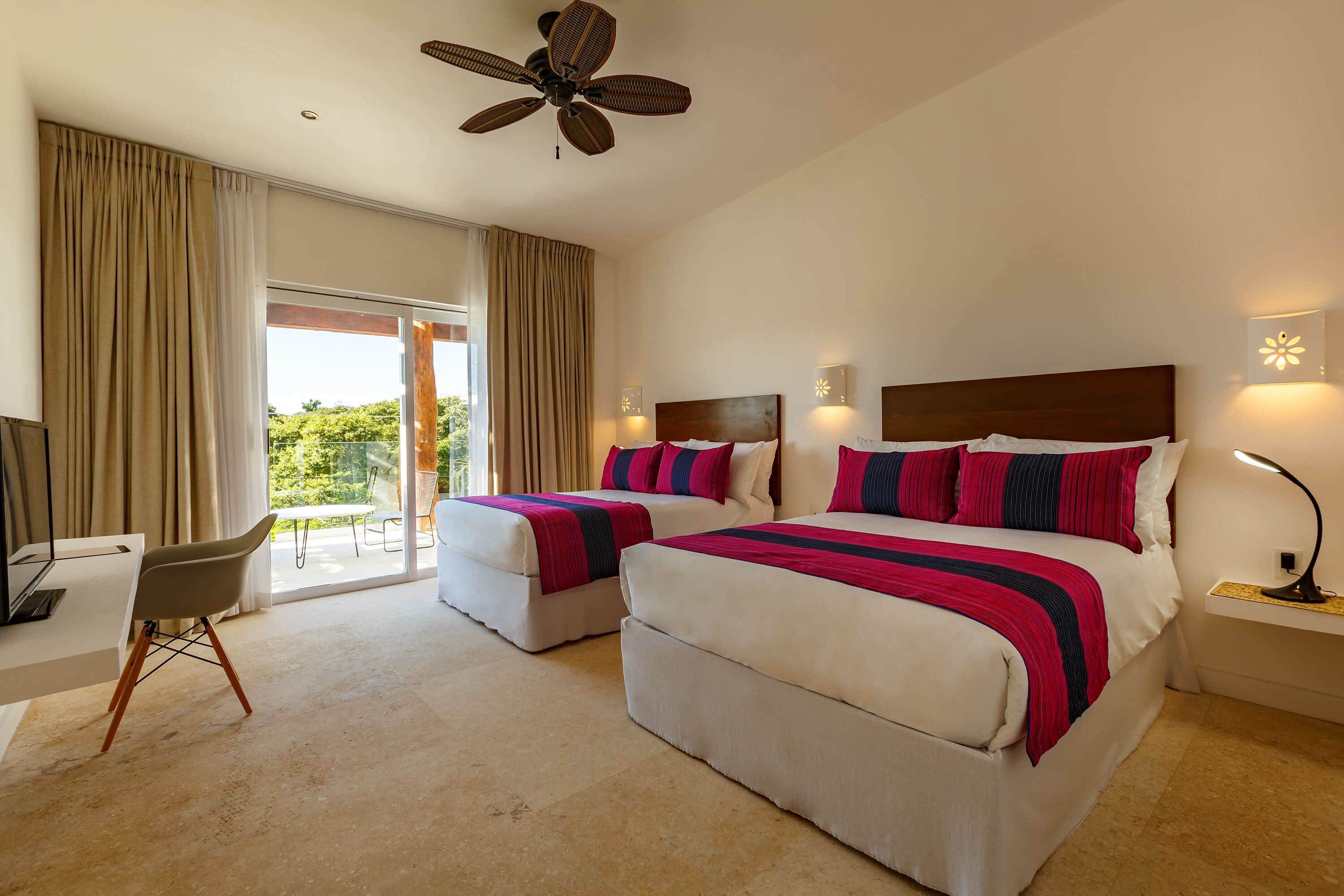 A hotel room with two beds, a desk, a chair, a ceiling fan, and a view of greenery outside.