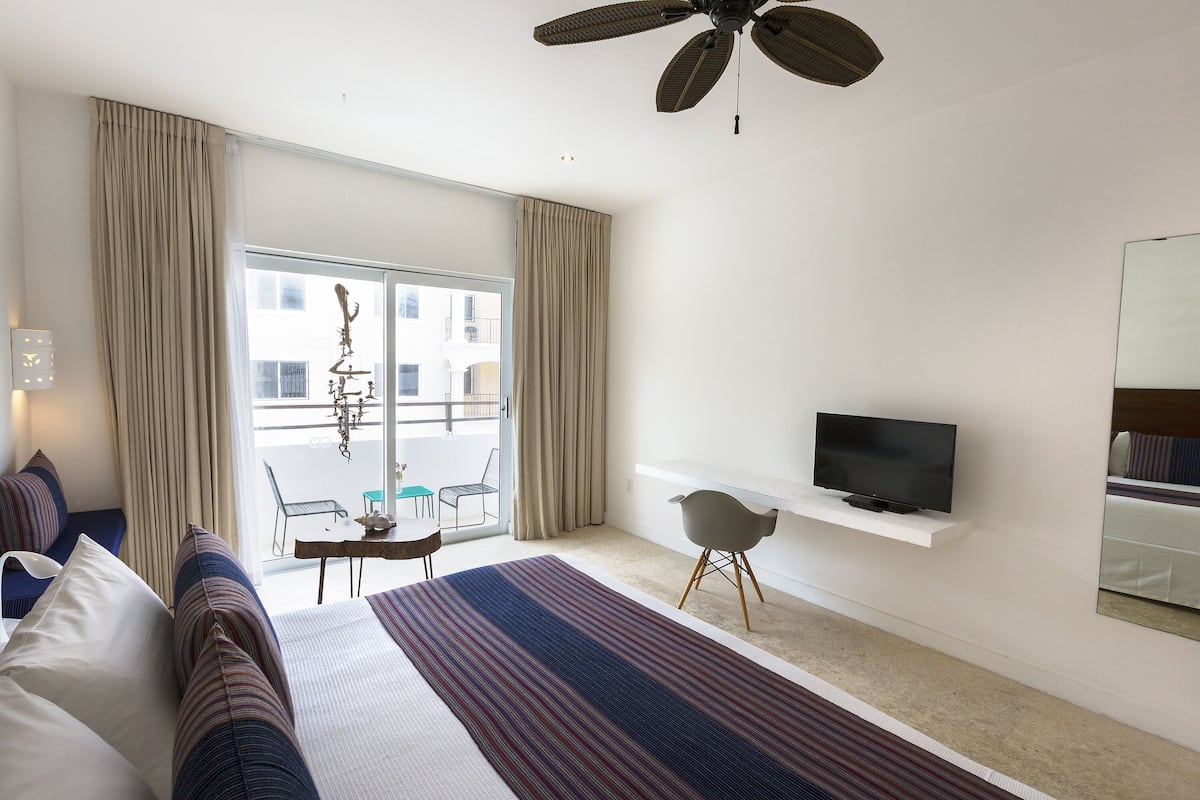 Ein Hotelzimmer mit Bett, Fernseher auf einem Tisch, Schreibtisch mit Stuhl und einem Balkon mit Vorhängen.