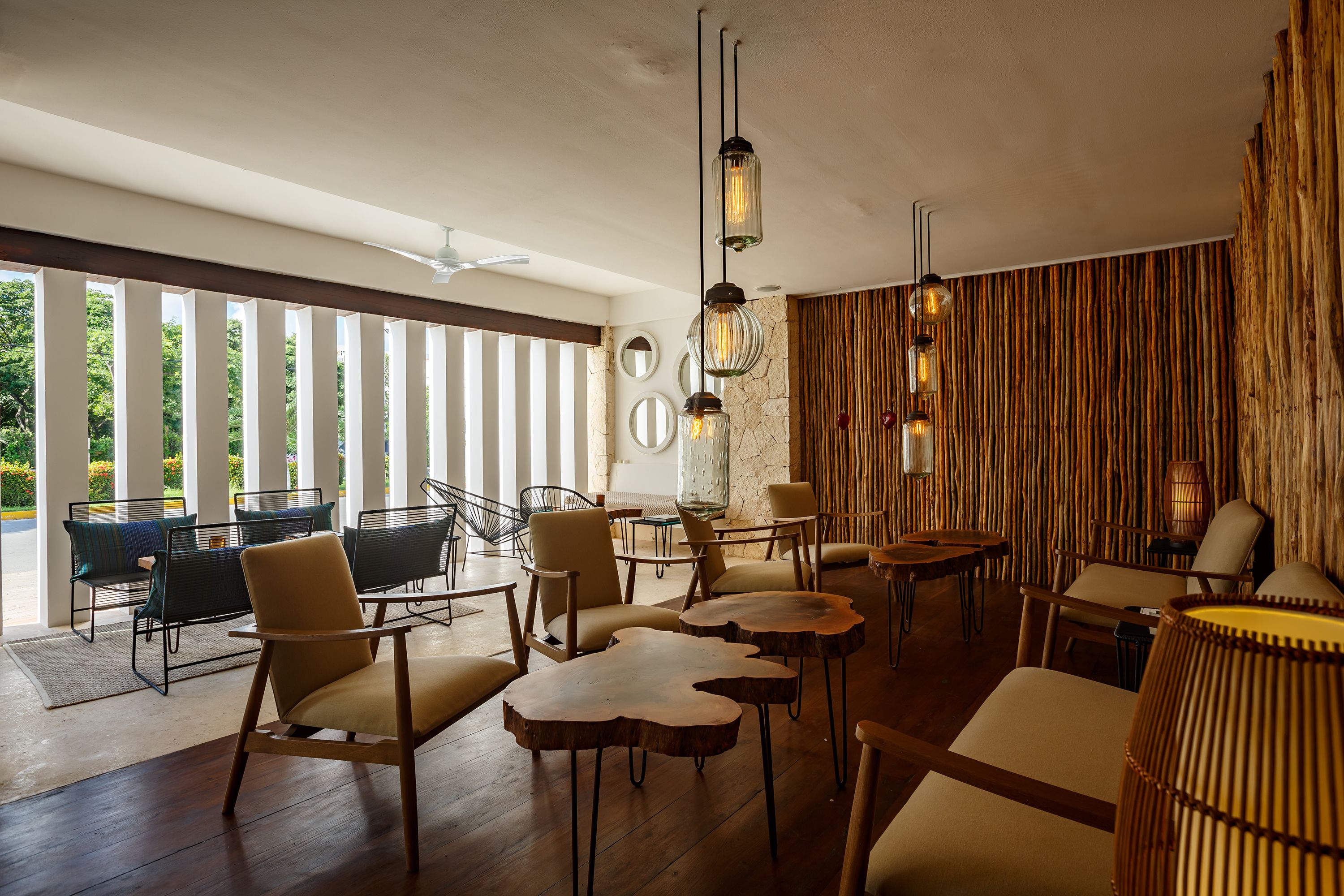 A modern lounge area with wooden furniture, pendant lighting, and a view of greenery through large windows.