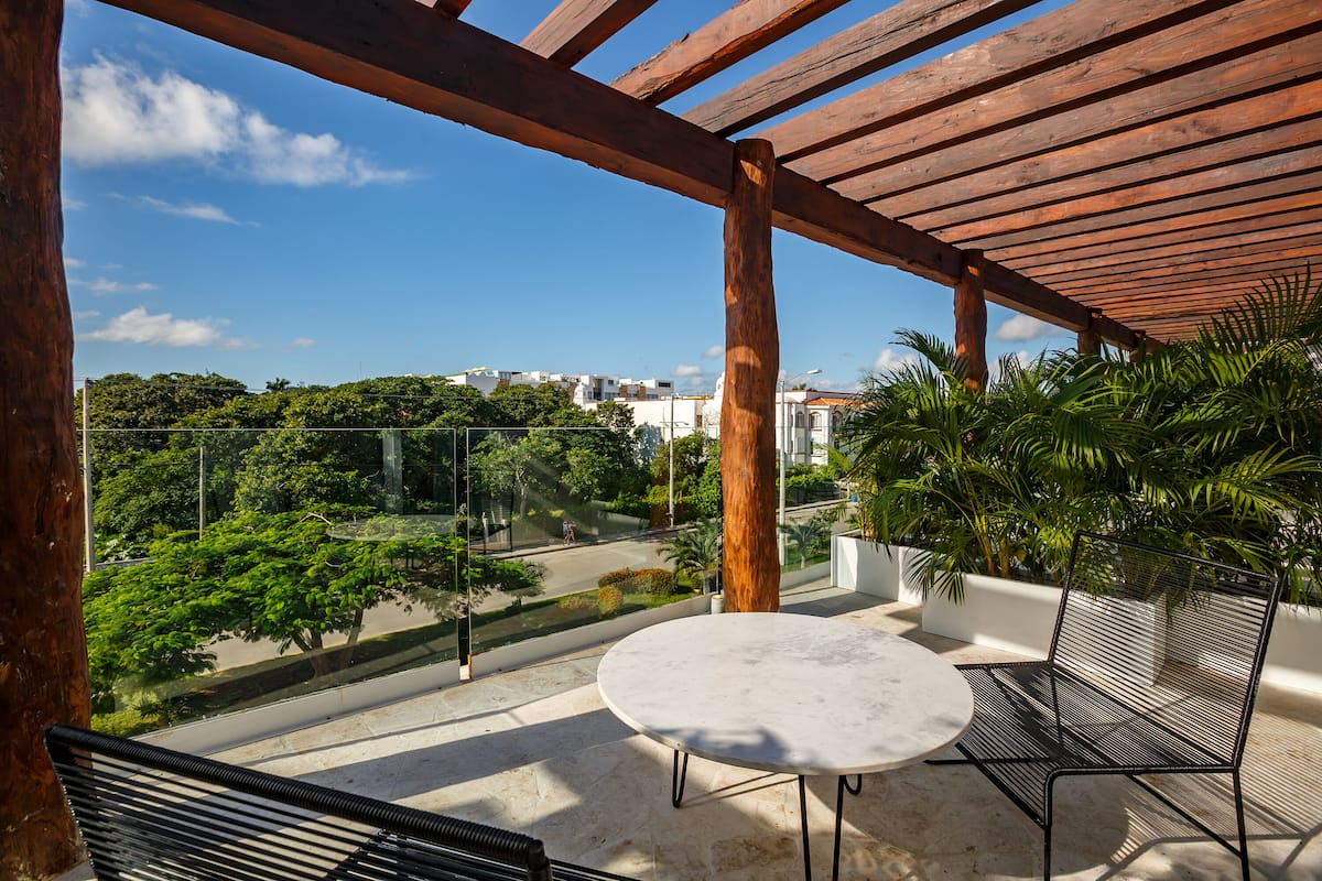 Ein Balkon mit weißem Tisch und Metallstühlen, mit Blick auf eine Stadtlandschaft mit Bäumen und Gebäuden.