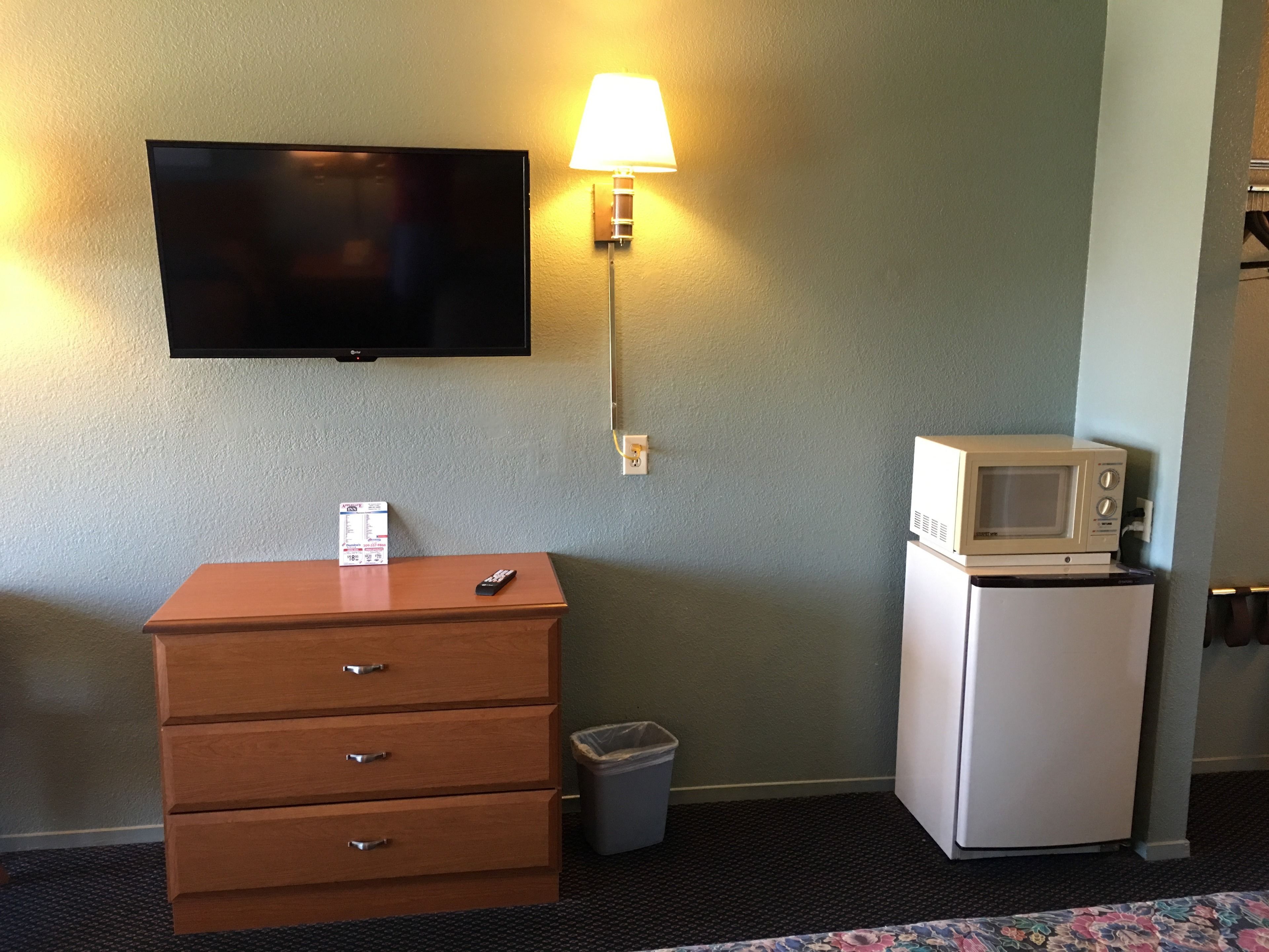 Una habitación de hotel con una televisión de pantalla plana montada en la pared, un pequeño aparador de madera con cajones, un cubo de basura y un microondas sobre una mesita.