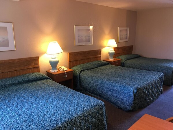 A hotel room with three single beds, each with a blue textured bedspread and a wooden headboard. There are bedside tables with lamps and a telephone. The walls are beige, and there are framed pictures above the beds.