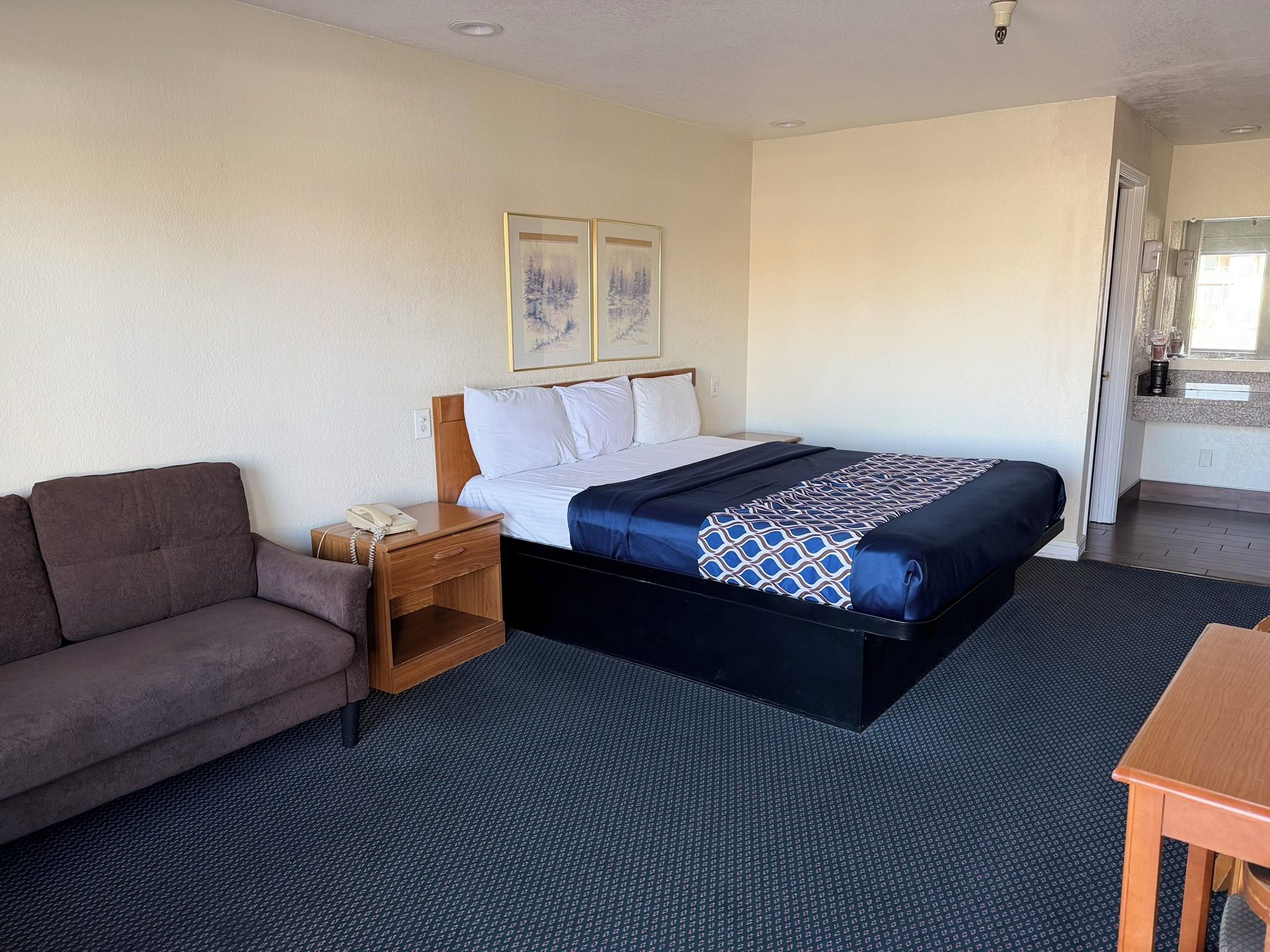 A hotel room with a bed, a sofa, a small table, a nightstand, and a kitchenette area.