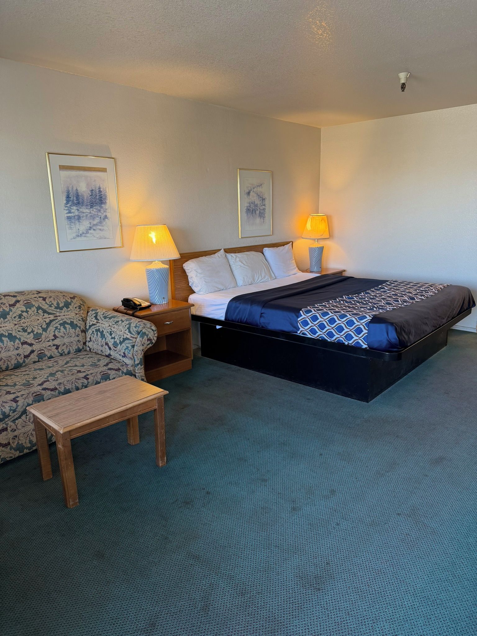 A hotel room with a bed, bedside lamps, a sofa, a small table, and two wall-mounted artworks.