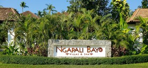 Señal de Ngapali Bay Villas & Spa rodeada de exuberante vegetación y edificios con techo de paja.