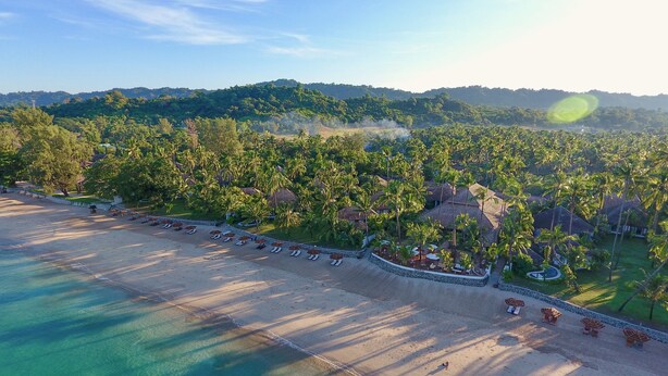 An aerial view of a beach resort with a long sandy beach, rows of sun loungers, and a backdrop of lush green hills.
