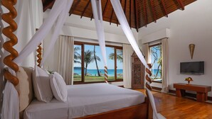 A bedroom with a wooden bed, white bedding, and a view of the beach and palm trees through large windows.