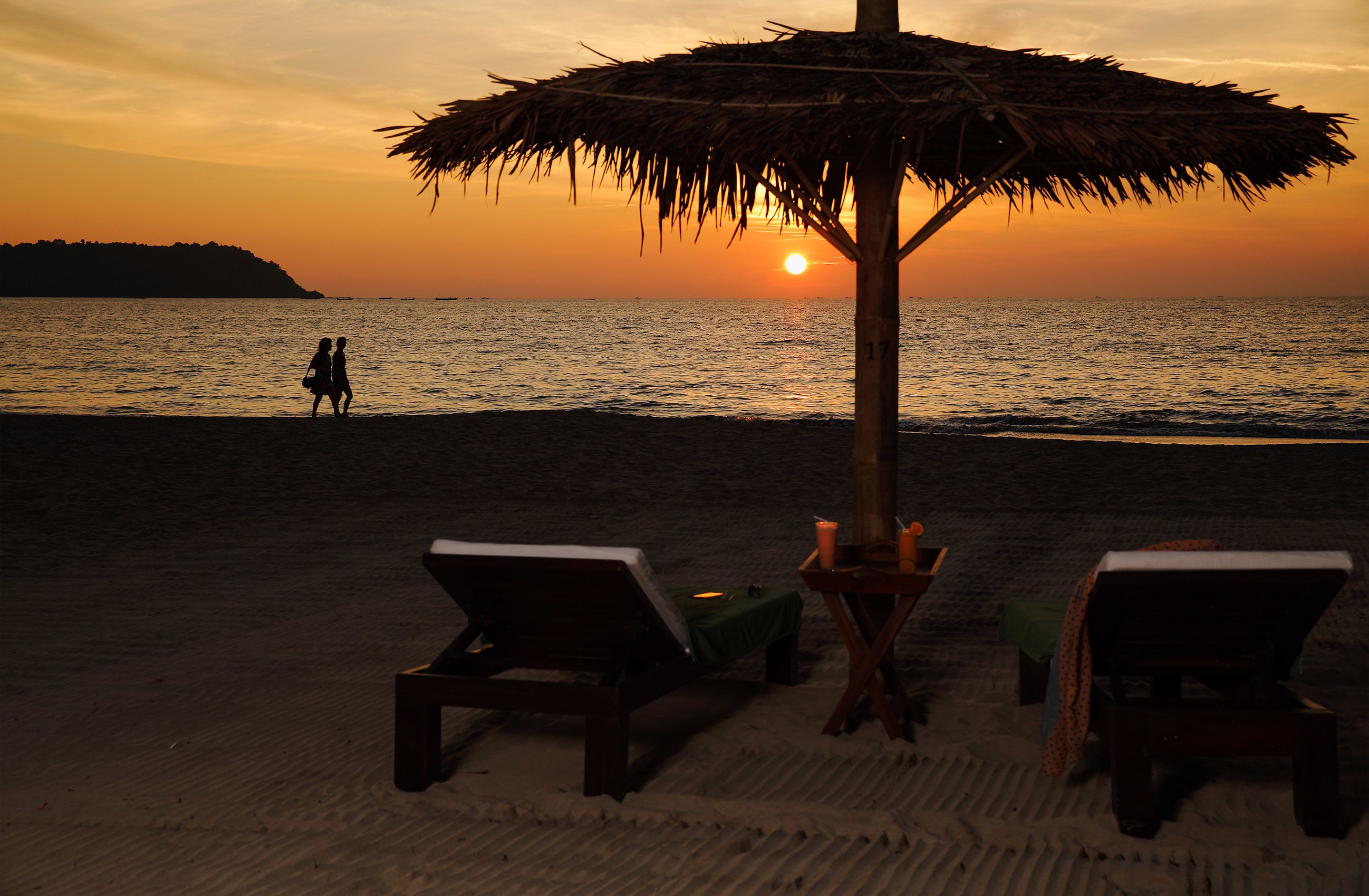 A beach scene with a sunset, two lounge chairs, a table with drinks, and a straw hat on the table.
