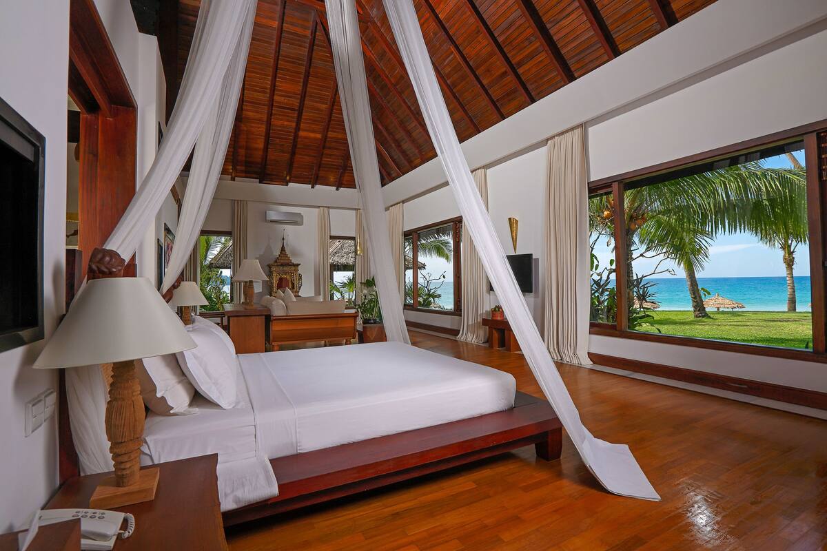 Een ruime slaapkamer met een groot bed, houten vloeren en uitzicht op de oceaan door grote ramen.