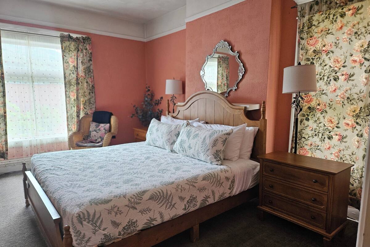 A bedroom with a wooden bed, a nightstand, a dresser, a chair, a mirror, and floral wallpaper.