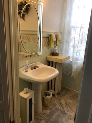 A bathroom with a pedestal sink, a mirror with a gold frame, a towel rack with towels, a trash bin, and a window with a floral curtain.