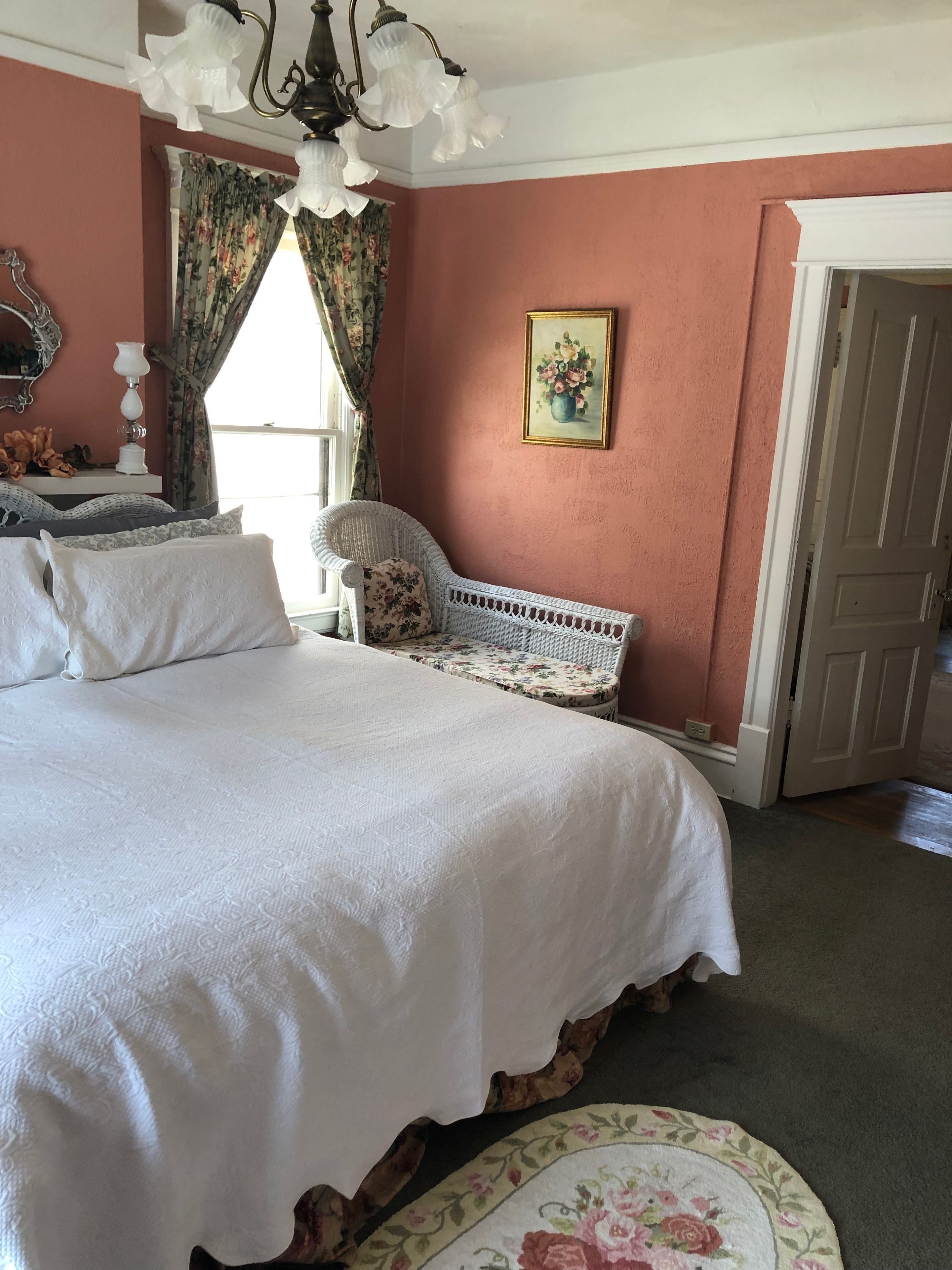 A bedroom with a large bed, a wicker chair, a painting, and a door.