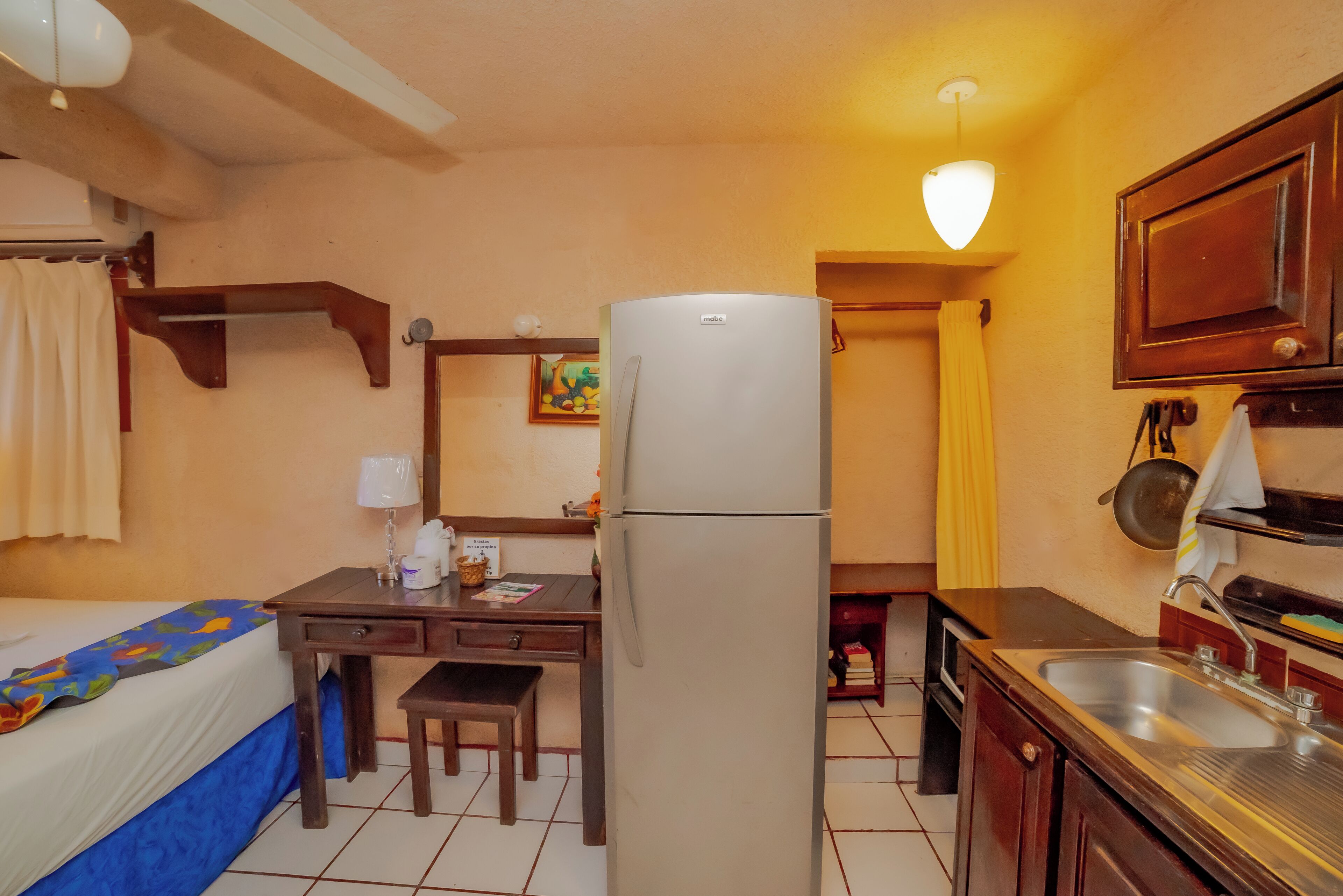 A compact hotel room with a bed, a desk with a chair, a refrigerator, a sink, and a small kitchenette.