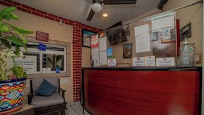 Zona de recepción con un escritorio rojo, una silla, una planta en maceta y un tablón de anuncios con diversos avisos.