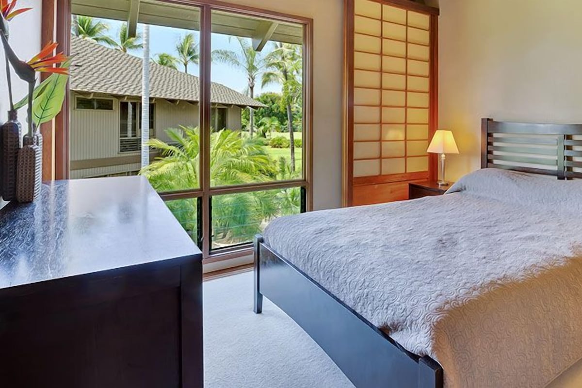 A bedroom with a bed, bedside table, lamp, and a view of a tropical landscape through a large window.