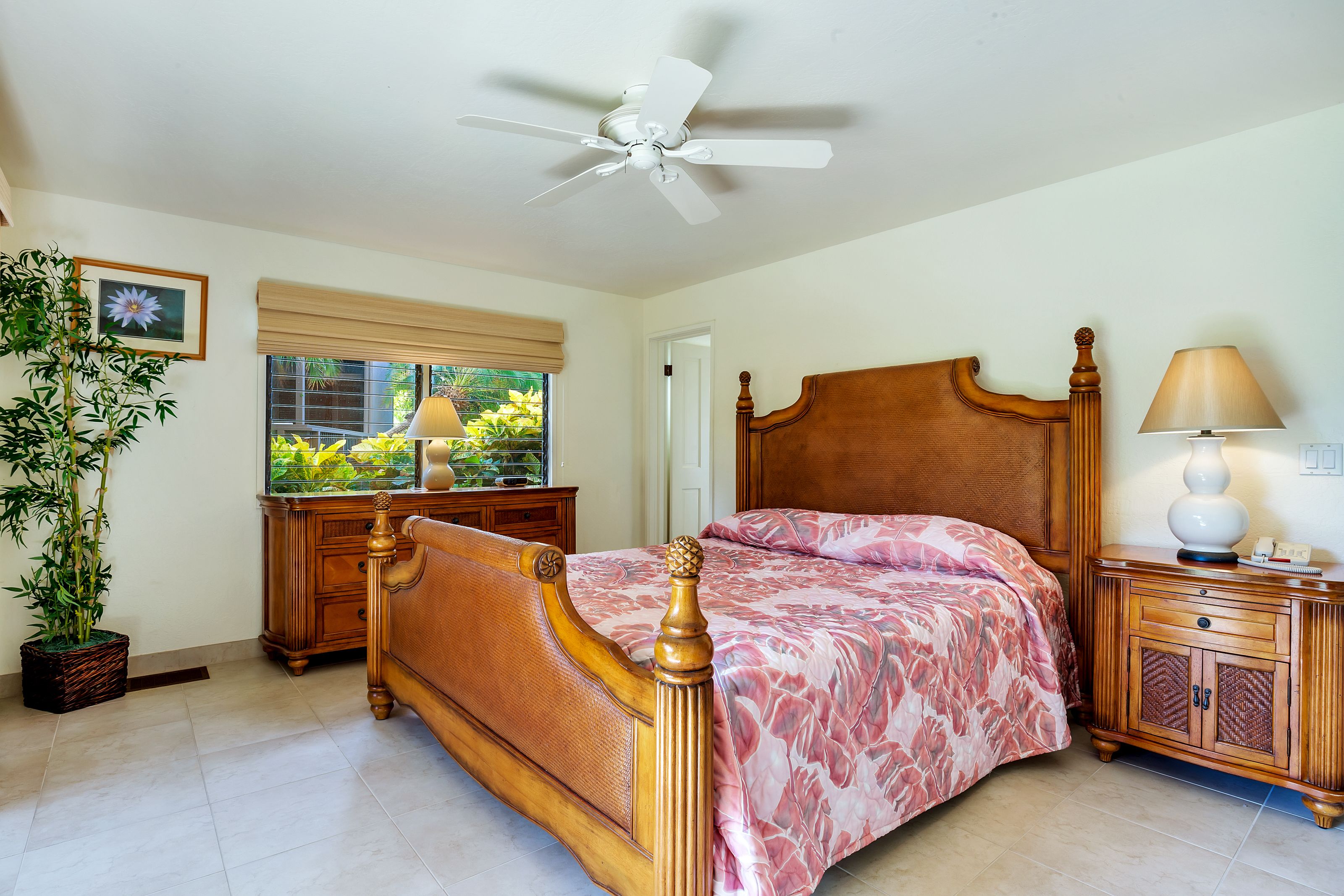 A bedroom with a wooden bed, a ceiling fan, a nightstand with a lamp, a window with blinds, and a potted plant.