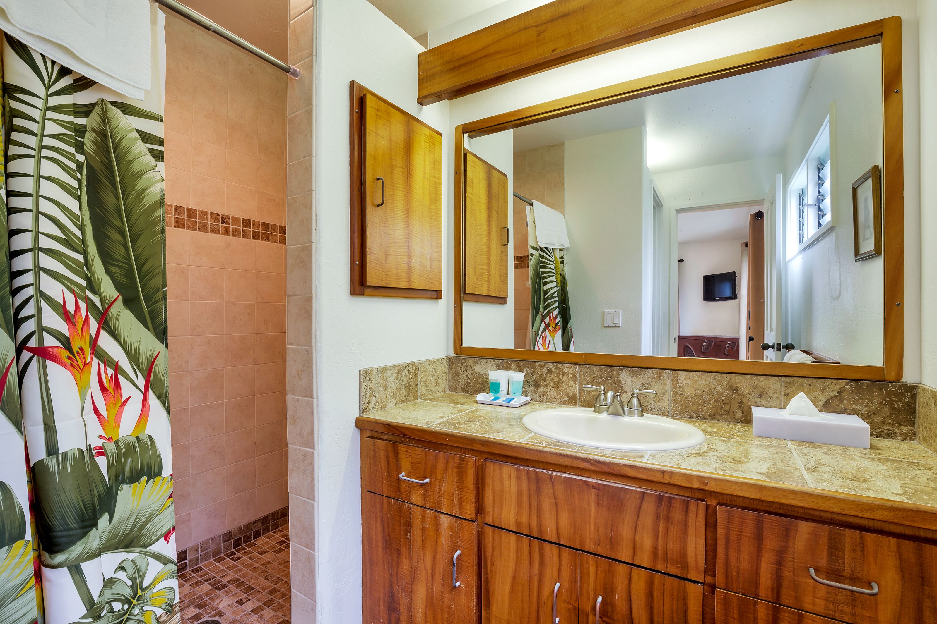 Une salle de bain avec un meuble-lavabo en bois, un grand miroir et une douche agrémentée d’un rideau de douche aux motifs tropicaux.