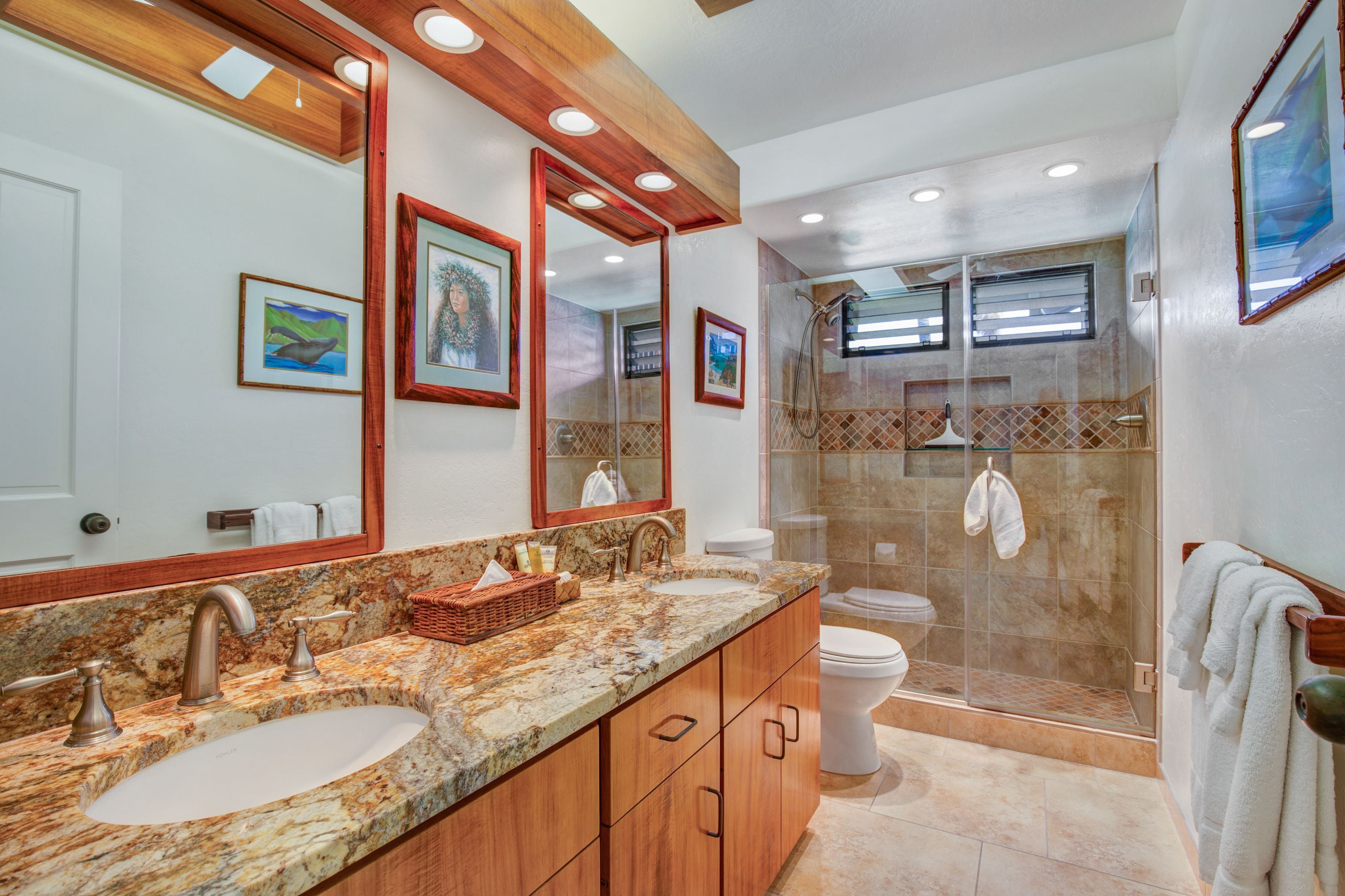 Une salle de bain avec un lavabo en marbre, un meuble-lavabo en bois, une cabine de douche en verre et des œuvres d’art encadrées accrochées aux murs.