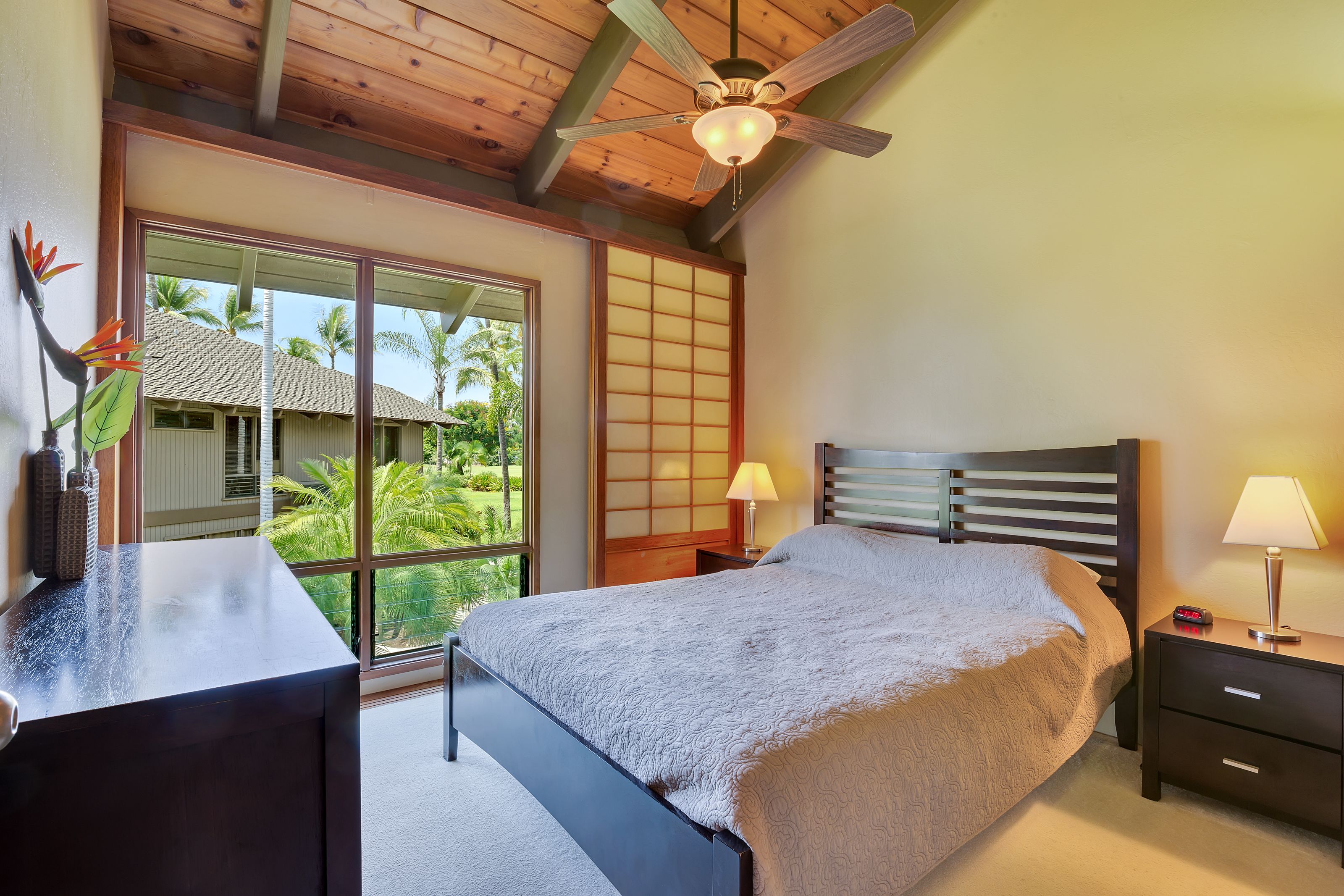 A bedroom with a bed, nightstands, a desk, a ceiling fan, and large windows with a view of palm trees.