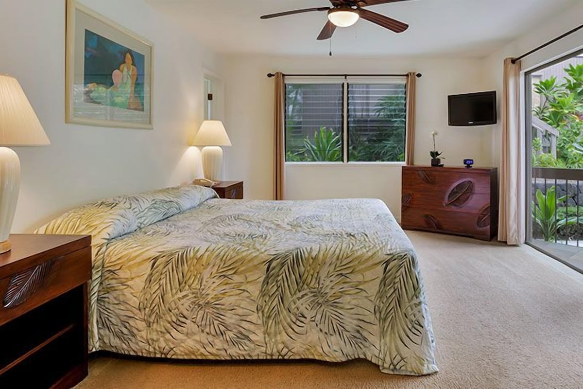 A bedroom with a bed, two bedside lamps, a ceiling fan, a window with blinds, a TV on the wall, and a view of a garden through the window.