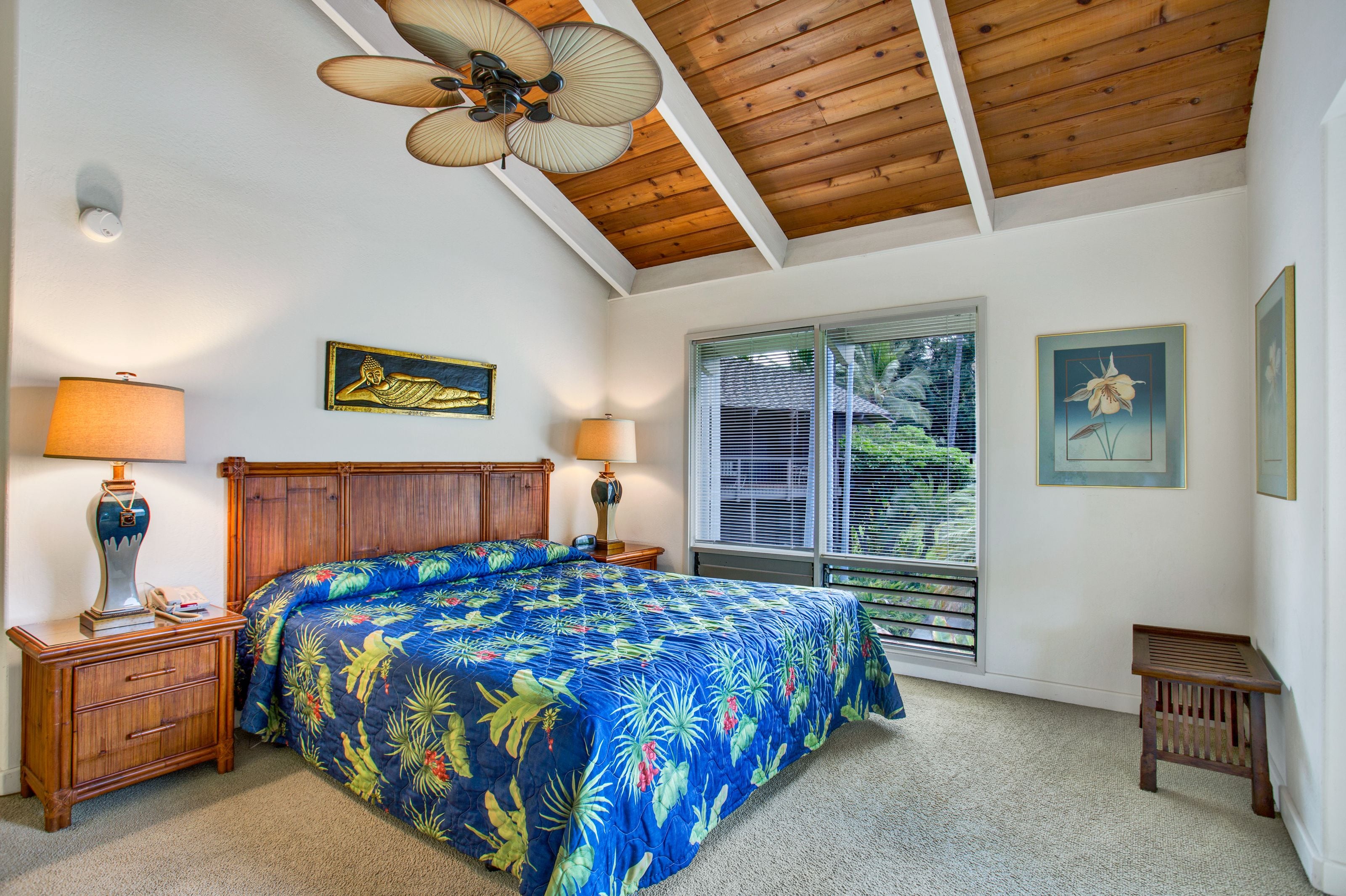 A bedroom with a wooden ceiling, a bed with a floral bedspread, two bedside lamps, a nightstand, a window with blinds, and a small wooden bench.