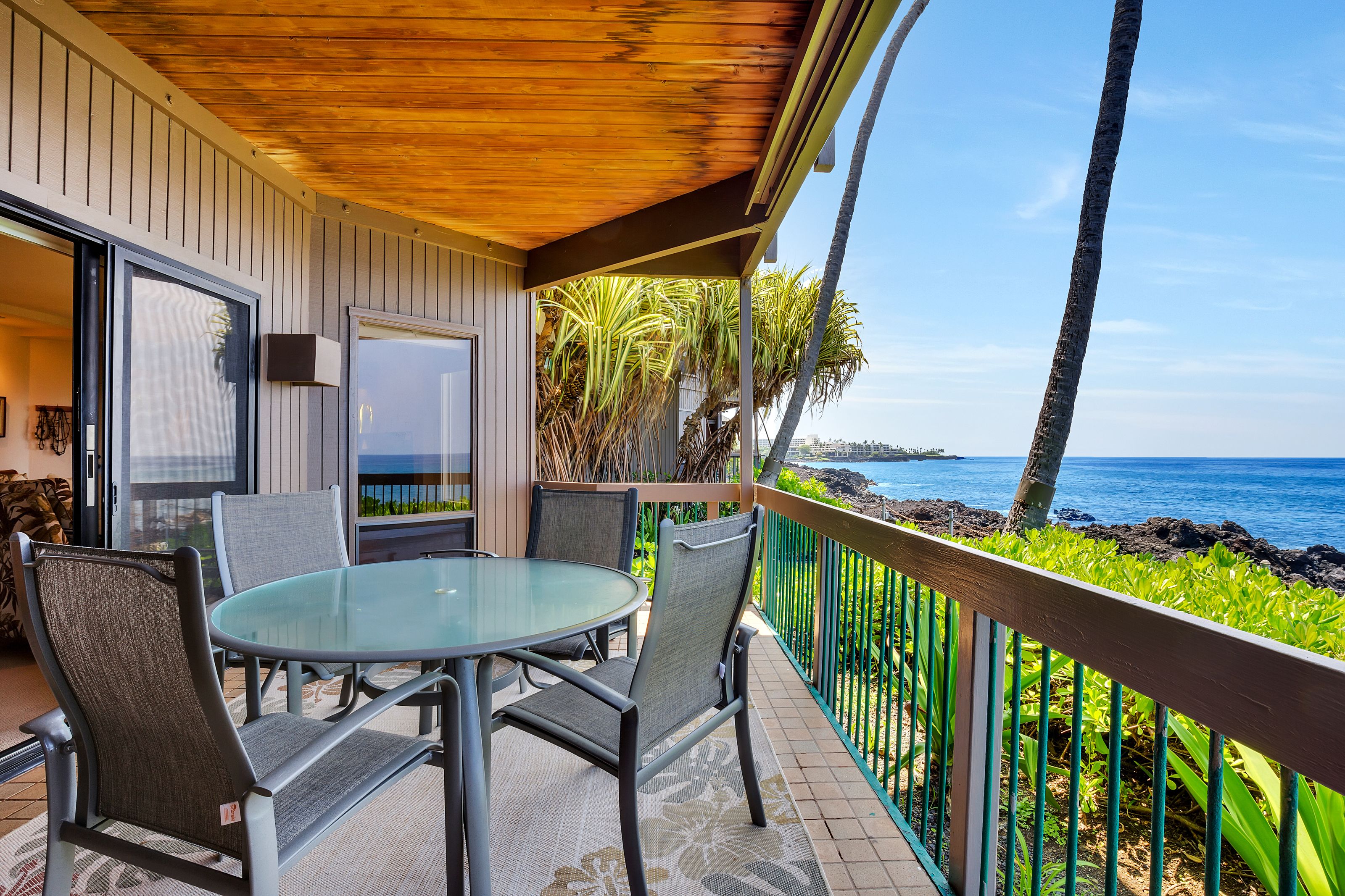 A covered outdoor seating area with a glass table and chairs, overlooking a rocky coastline and the ocean.