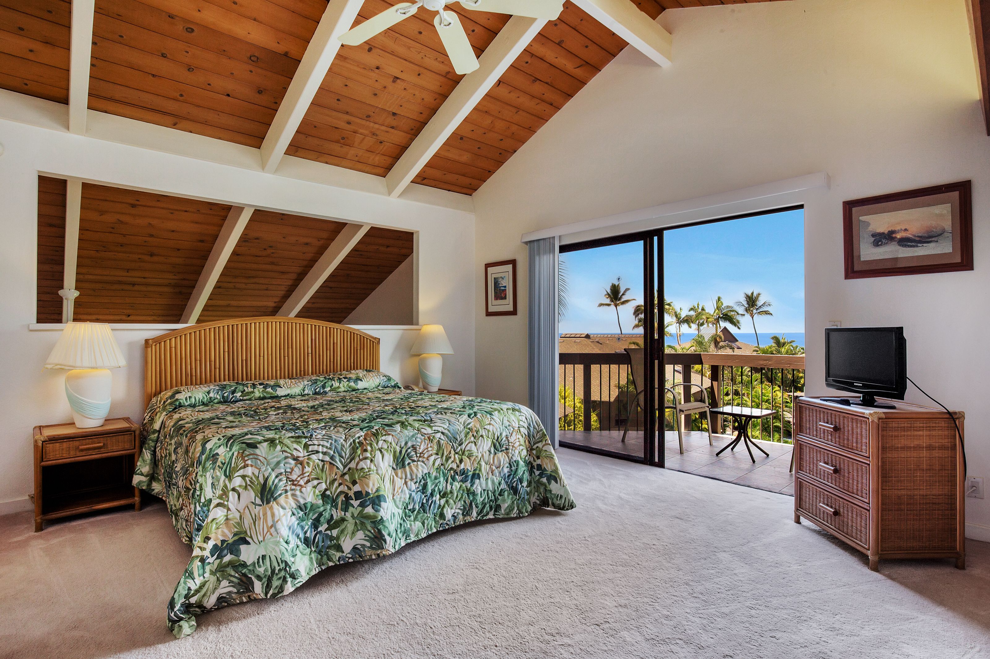 A bedroom with a large bed, wooden ceiling beams, a balcony with a view of palm trees, and a television on a cabinet.