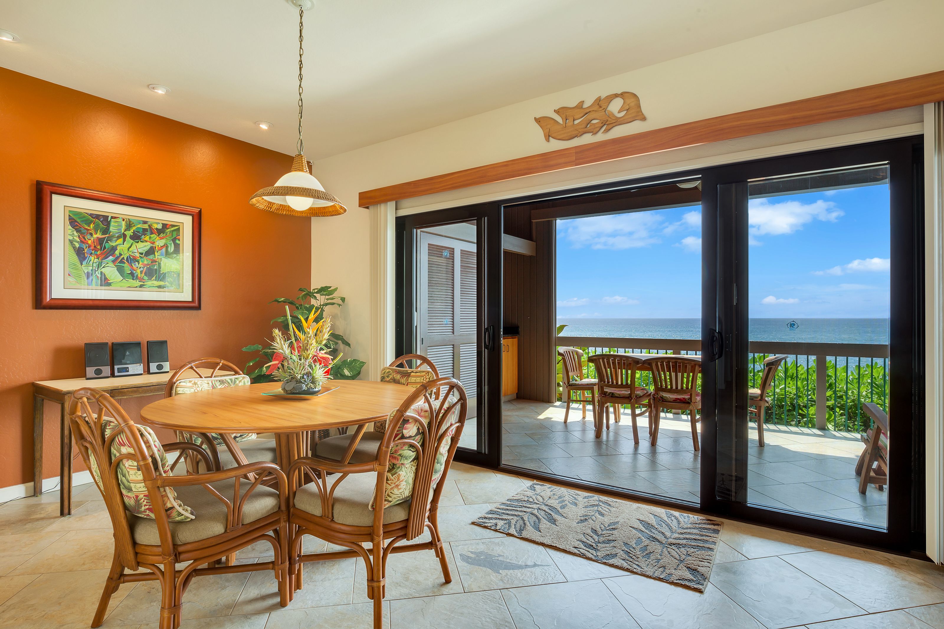 A dining area with a round table and chairs, a framed artwork on the wall, and a view of the ocean through a sliding glass door.