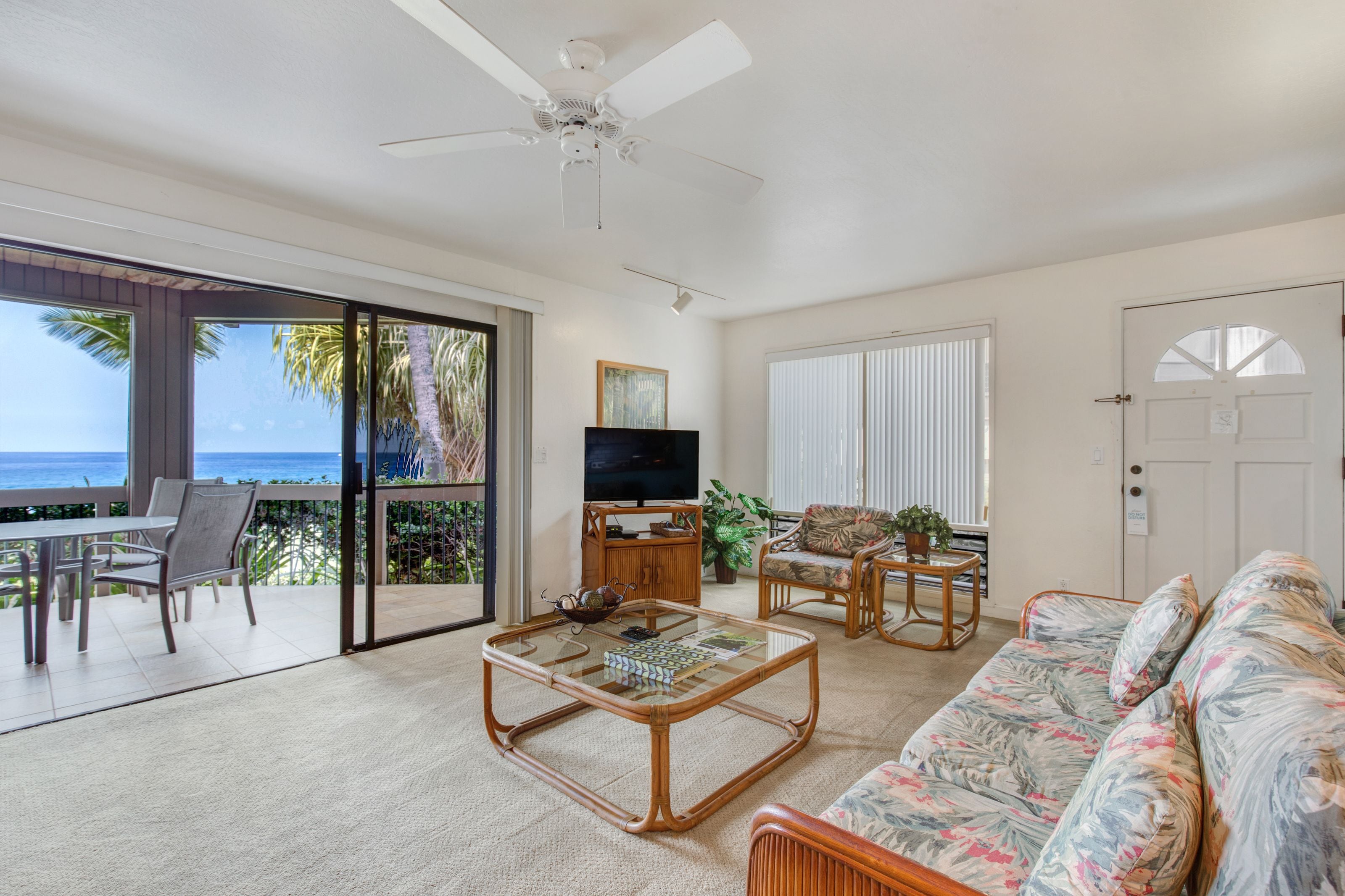 A spacious living room with a large glass door leading to a balcony, a ceiling fan, a flat-screen TV, a coffee table, and a sofa.