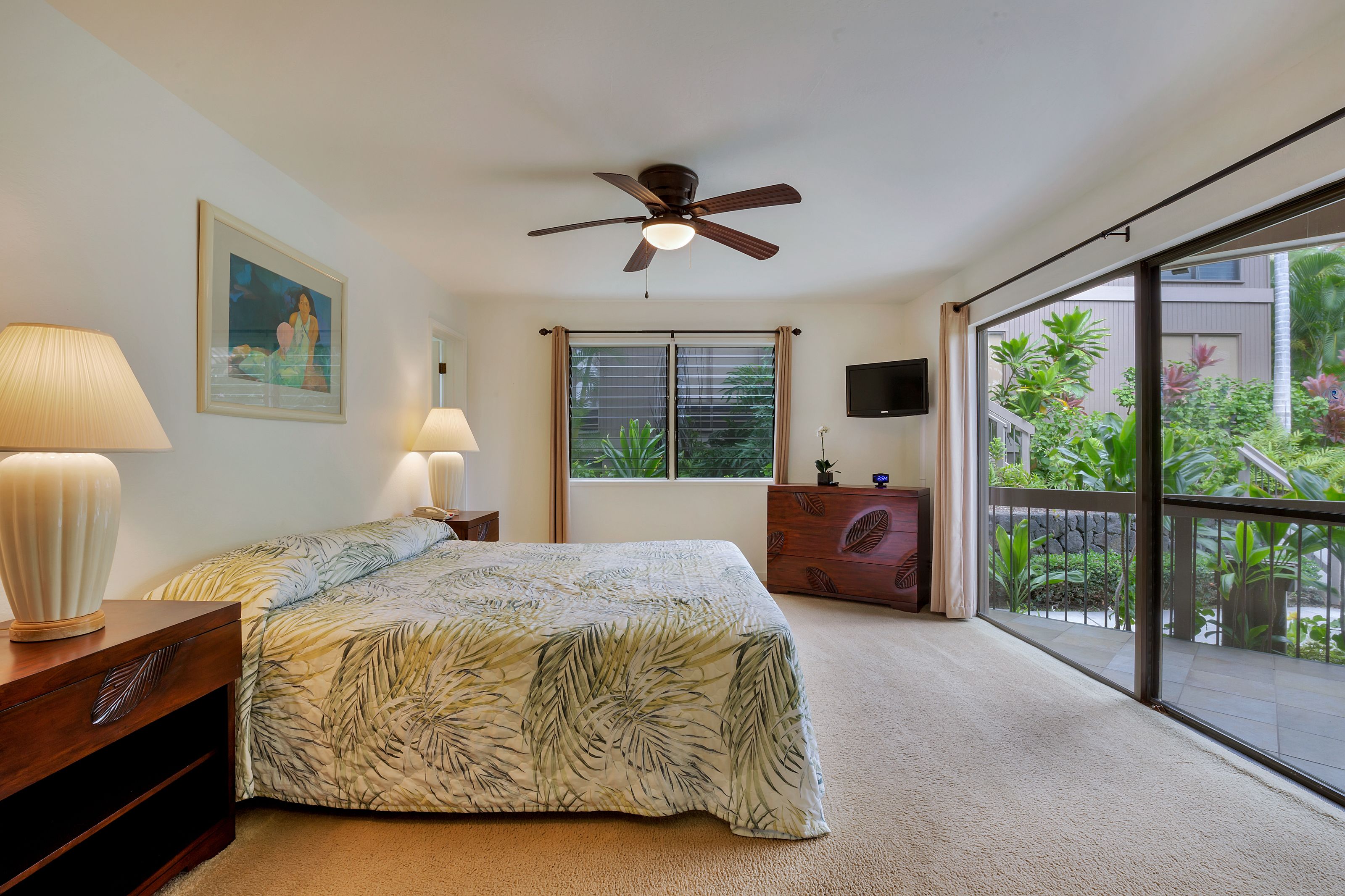 Une chambre à coucher avec un lit, un ventilateur de plafond, une fenêtre avec des rideaux, un téléviseur et une vue sur la verdure à l’extérieur.