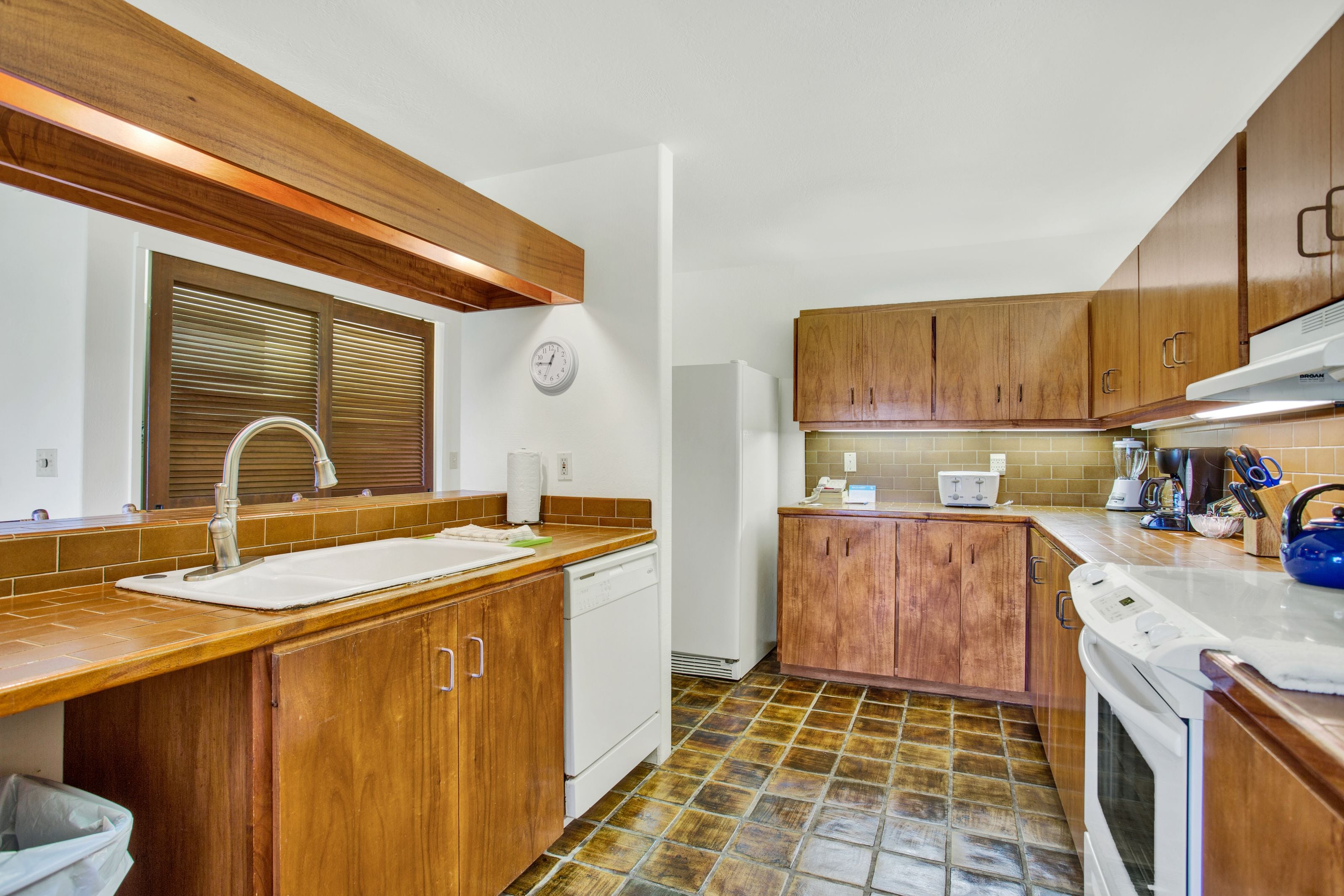 Une cuisine avec des meubles de rangement en bois, un évier, un lave-vaisselle, une cuisinière et une horloge au mur.