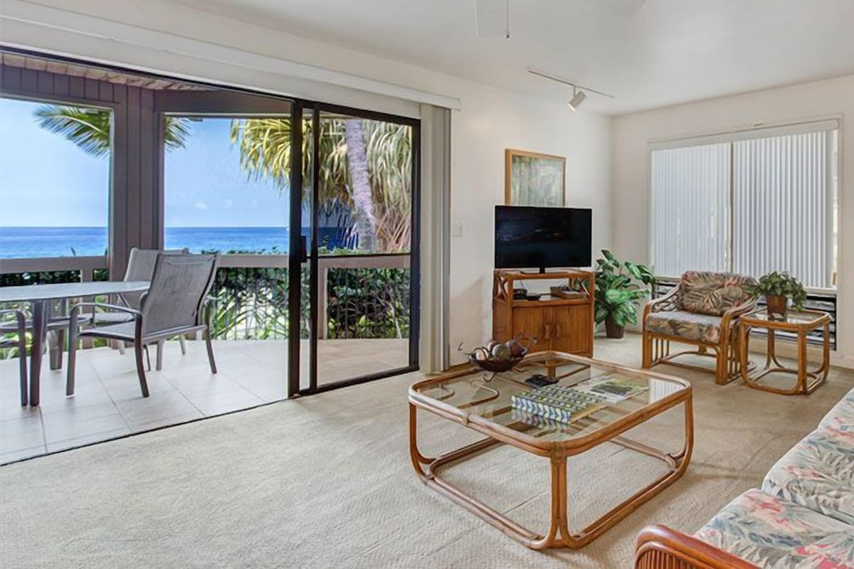 A spacious living room with a glass coffee table, a sofa, and a TV. There is a balcony with a view of the ocean.