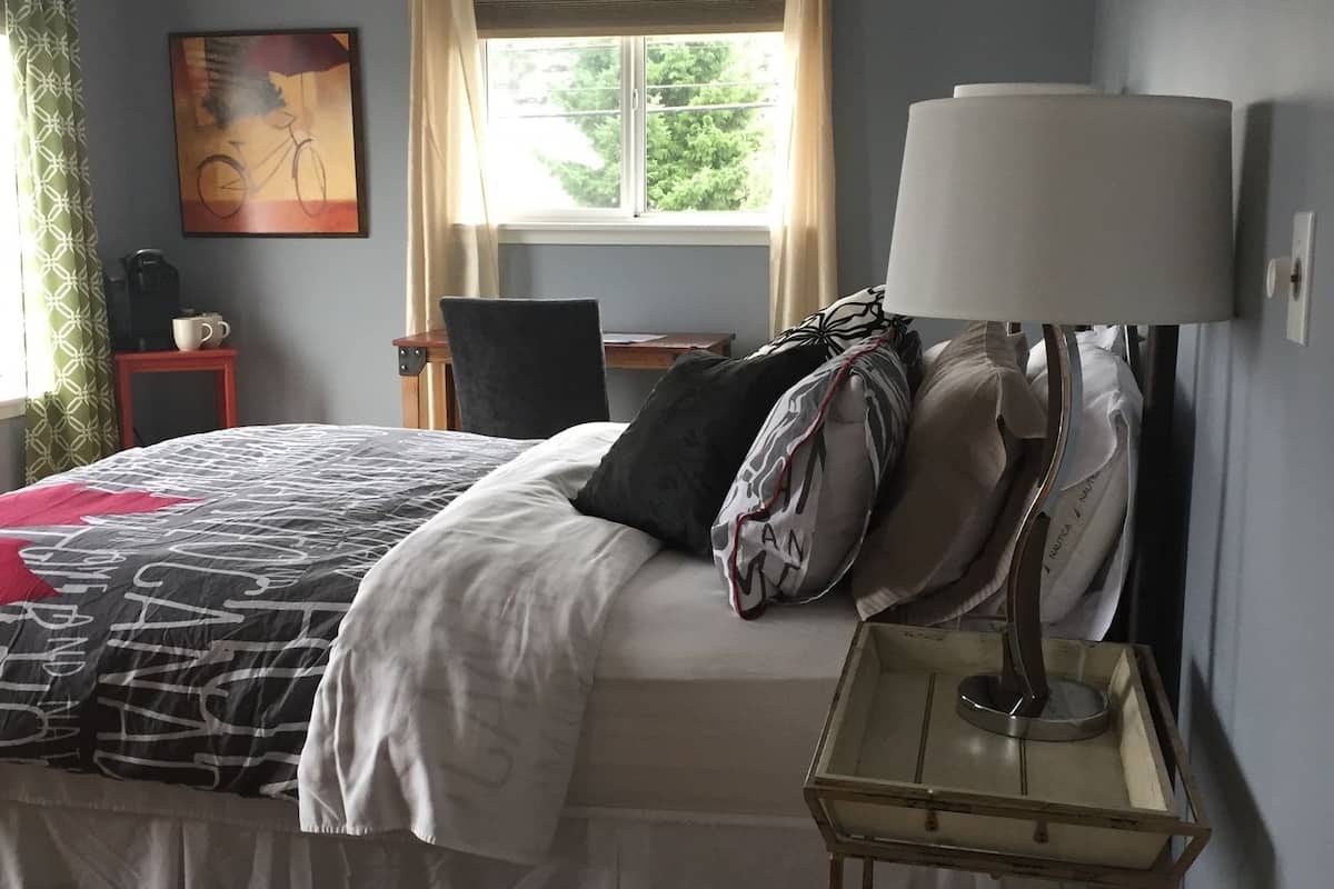 Un dormitorio con cama, una mesita de noche con lámpara, una silla, una pequeña mesa con cafetera y una ventana con cortinas. En la pared, un cuadro.