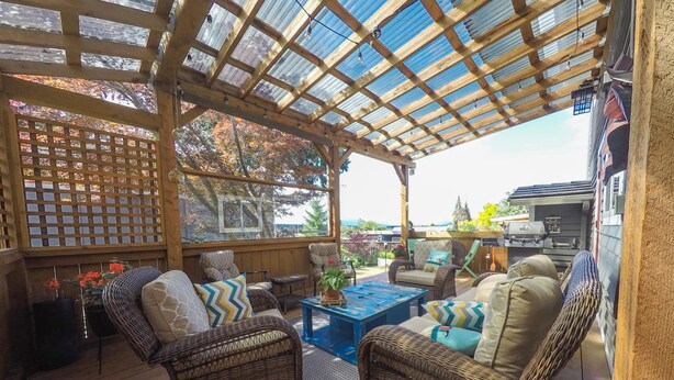 Un área de estar techada al aire libre con mobiliario de mimbre y una mesa azul, con vistas a un patio trasero que tiene una parrilla y una barbacoa.