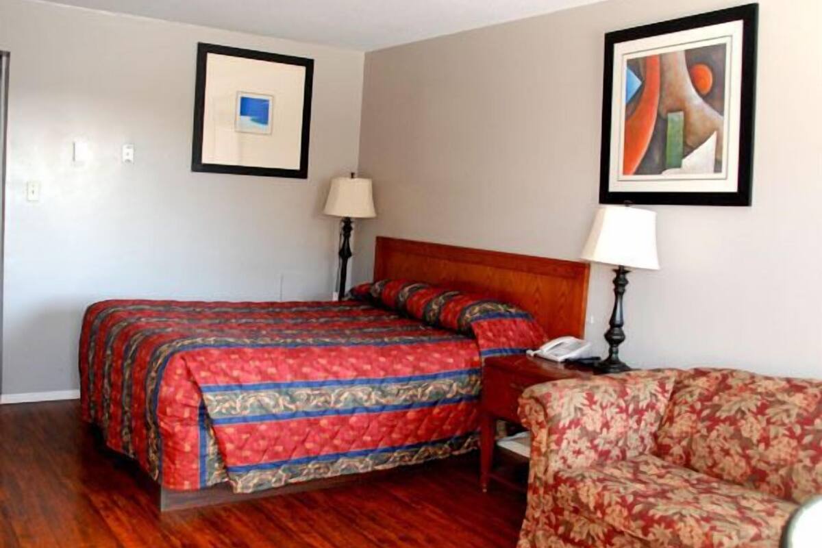 Quarto de hotel com uma cama, uma cadeira, uma mesinha de cabeceira, um abajur e duas molduras na parede.