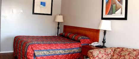 Habitación de hotel con cama, una silla, una mesita de noche, una lámpara y dos cuadros en la pared.