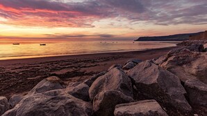 Una playa con rocas grandes al frente y un atardecer en el fondo.