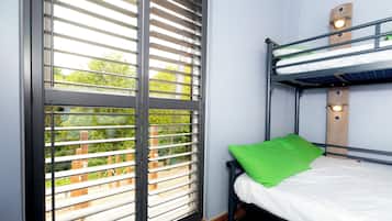 Una cama individual con almohada verde, una ventana con persianas y vistas al exterior con vegetación.