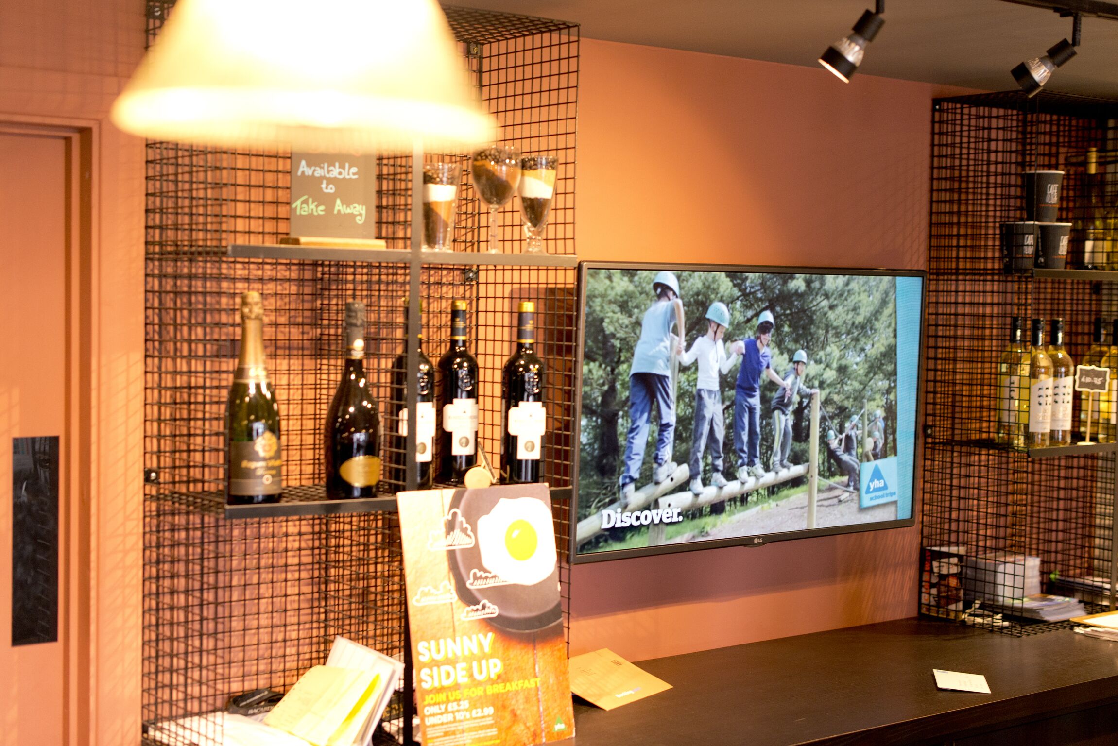 A bar area with a television displaying an outdoor scene, shelves with bottles, and a sign advertising a breakfast option.