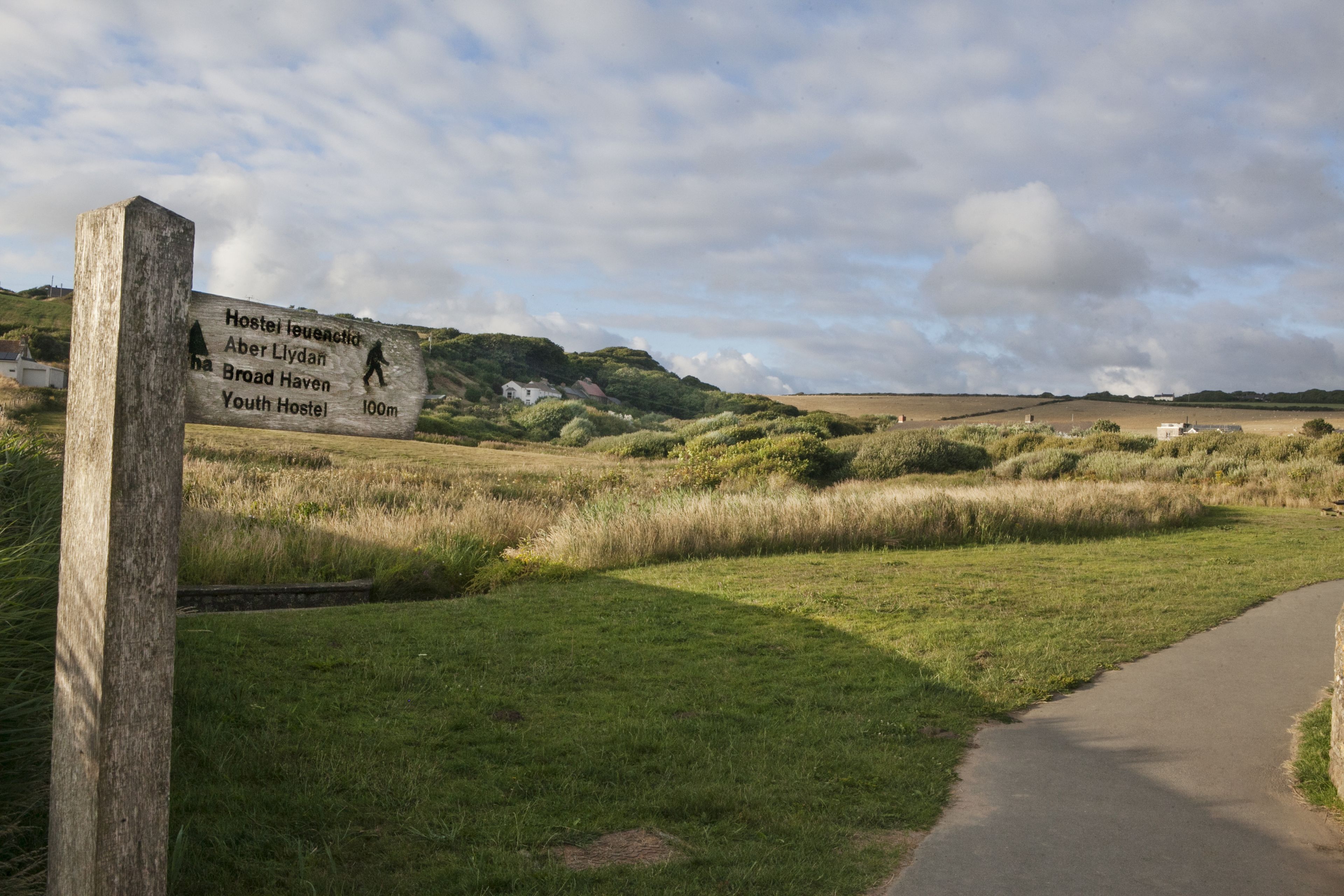 A signpost indicating directions to Hostel Ieuenctic, Aber Llydan, Broad Haven, and Youth Hostel, with a path leading through a grassy area.