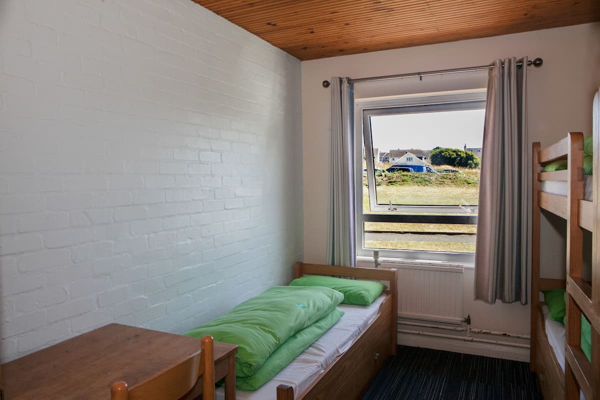 A small, single-bed room with a wooden desk and a window with curtains.