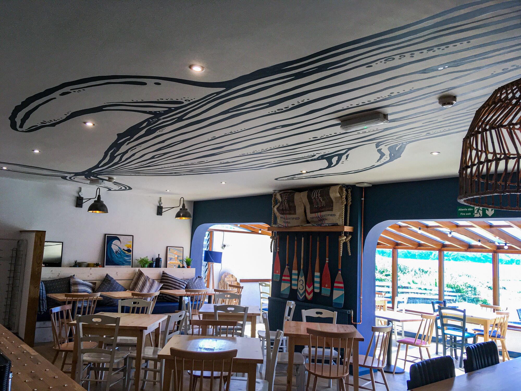 Ein Restaurantinneres mit Holztischen und -stühlen, einem Wandgemälde an der Decke und Blick ins Freie durch große Fenster.